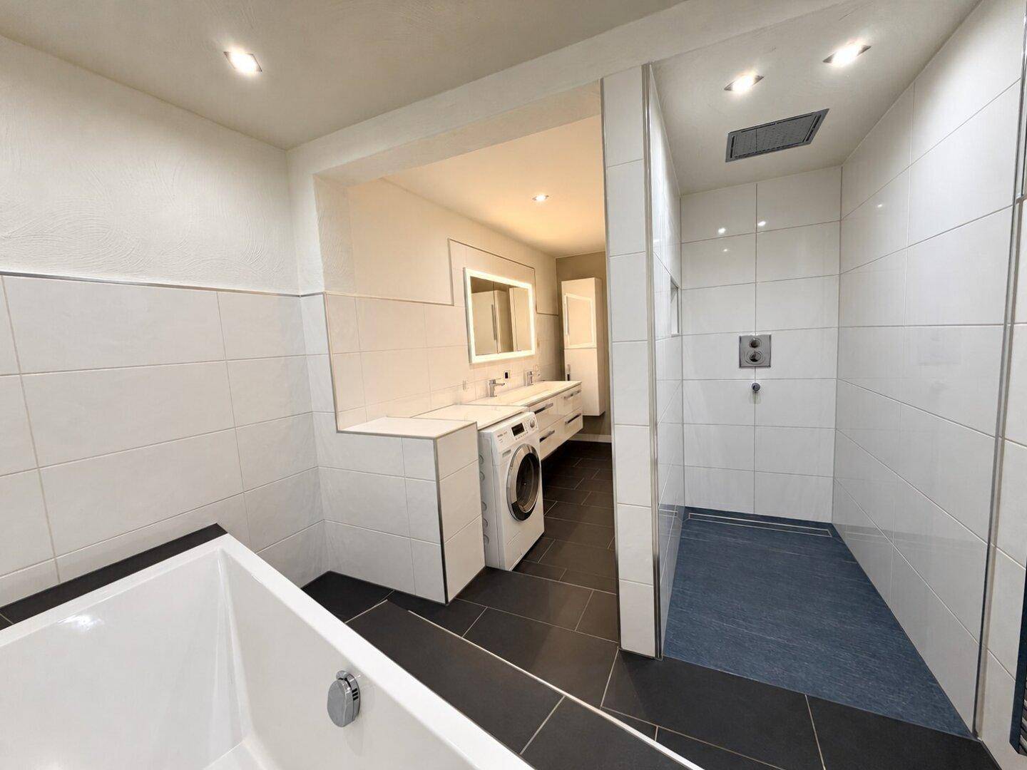 Modernes Badezimmer mit Badewanne, begehbarer Dusche, Waschmaschine und weißen Fliesen.