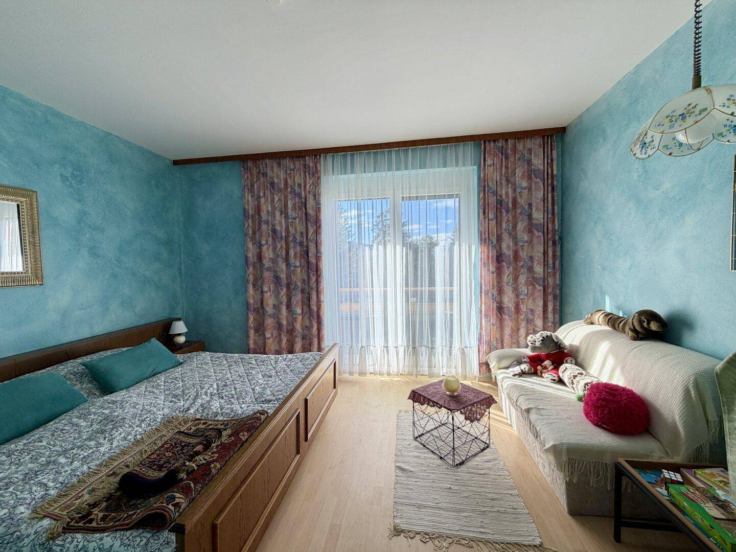 Schlafzimmer mit blauen Wänden, Doppelbett und hellem Fenster mit Vorhängen.