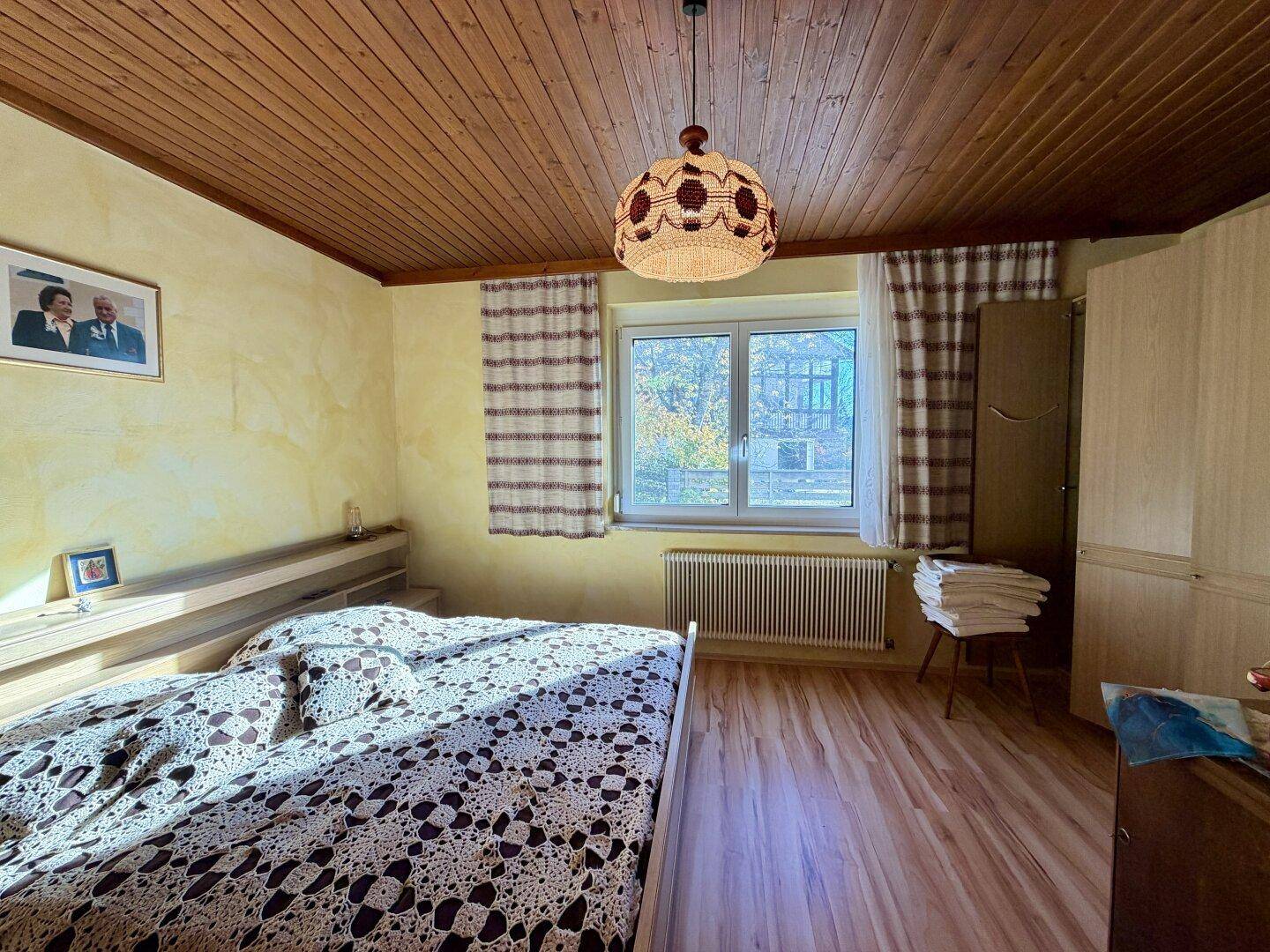 Schlafzimmer mit gelben Wänden, Holzbalkendecke und großem Fenster mit Blick ins Grüne.