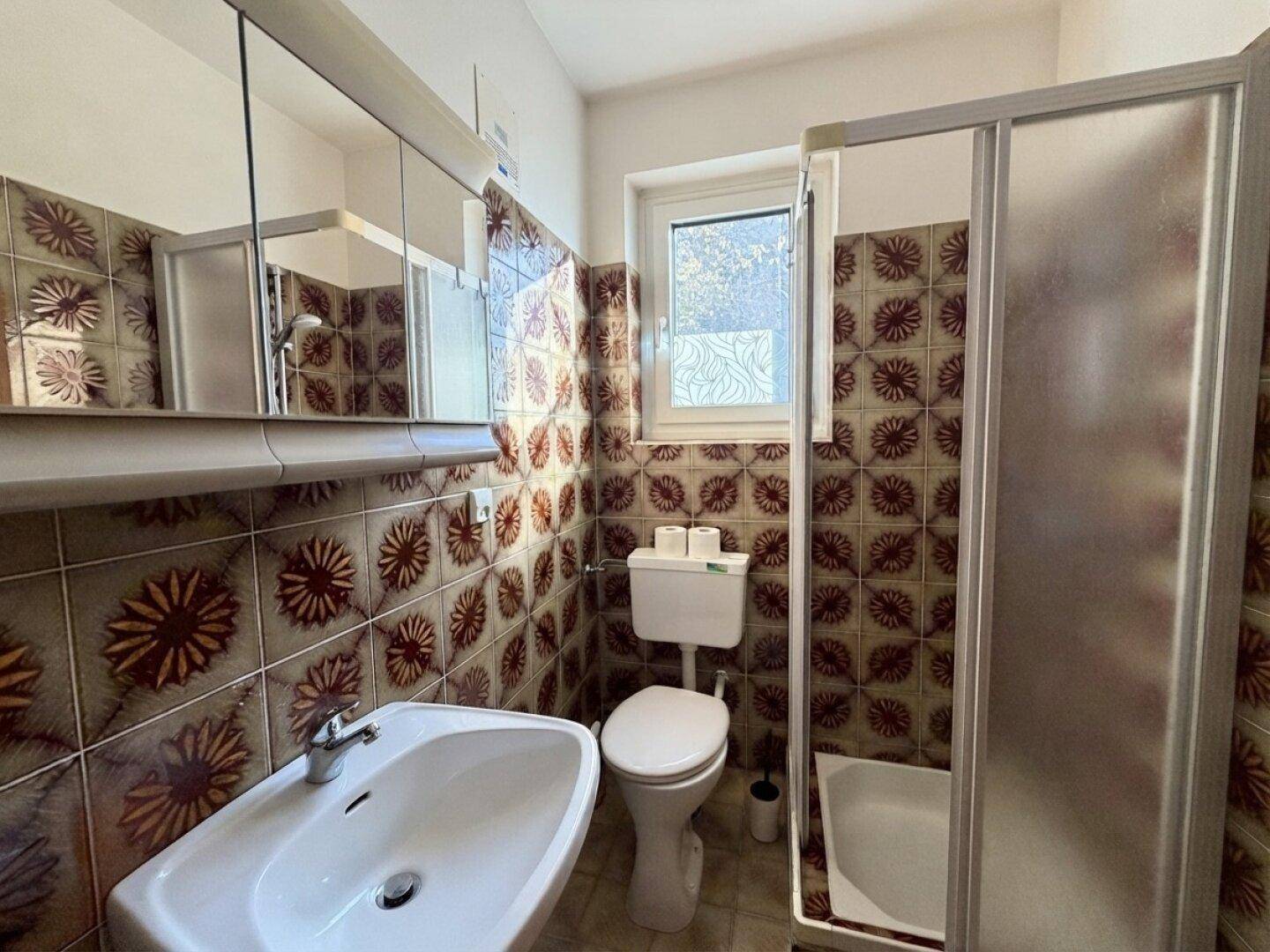 Badezimmer mit gemusterten Fliesen, Dusche, WC und Fenster für Tageslicht.
