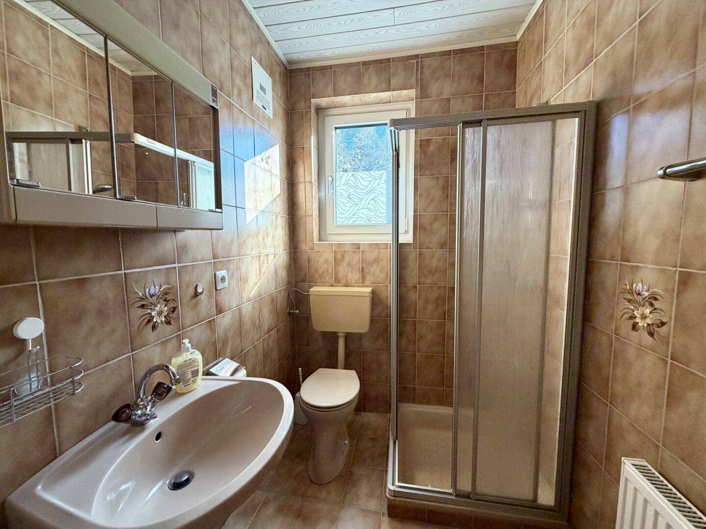 Badezimmer mit braunen Fliesen, Dusche, WC und Fenster für natürliche Belüftung.