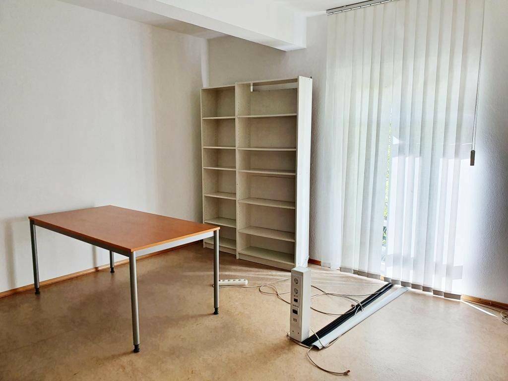 Helles Büro mit Tisch, weißem Regal und Fenster mit vertikalen Jalousien, ideal für konzentriertes Arbeiten.