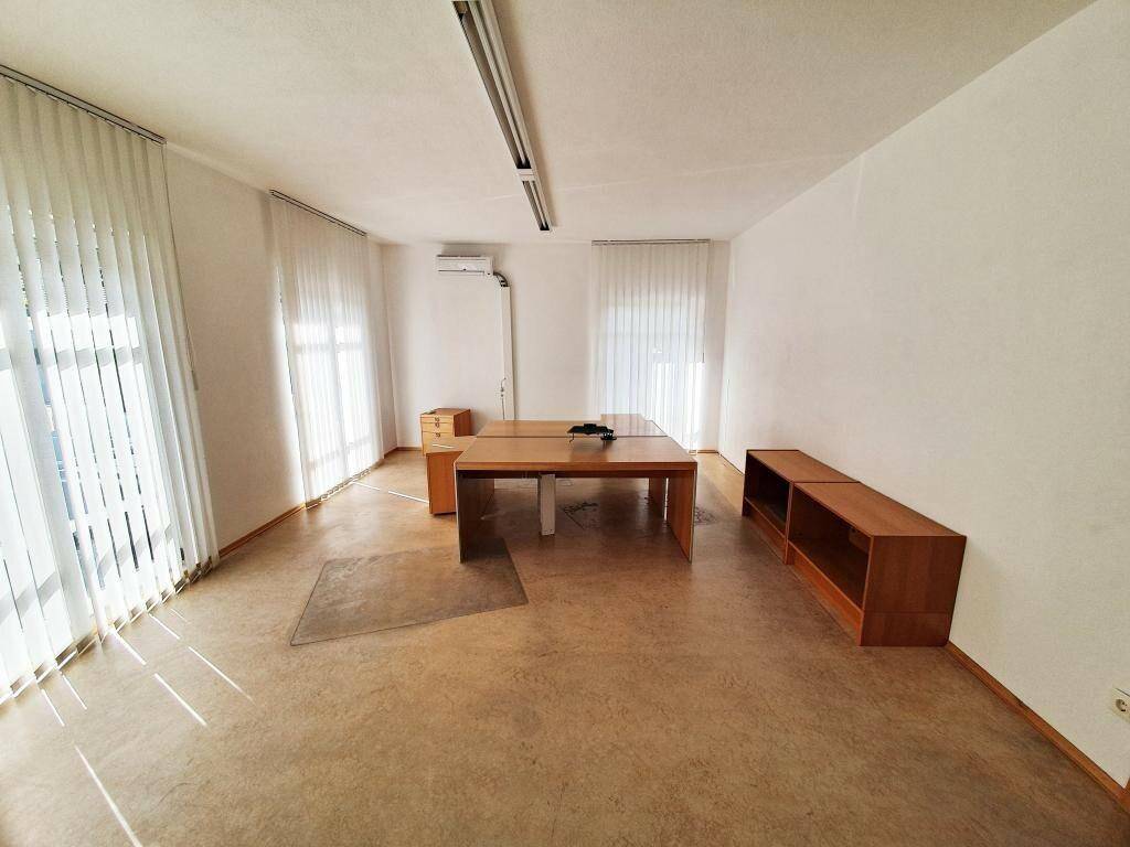 Geräumiges Büro mit Linoleumboden, großen Fenstern mit Jalousien, Holztisch und Klimaanlage.