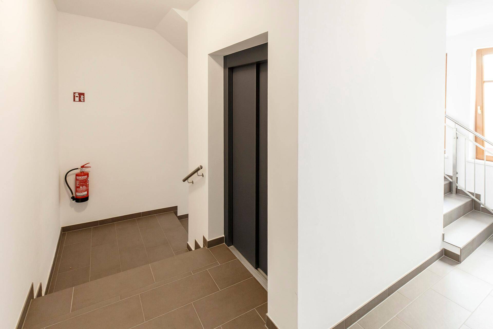 Treppenhaus mit modernem Aufzug, gefliesten Stufen und einem Feuerlöscher an der Wand, gut beleuchtet.