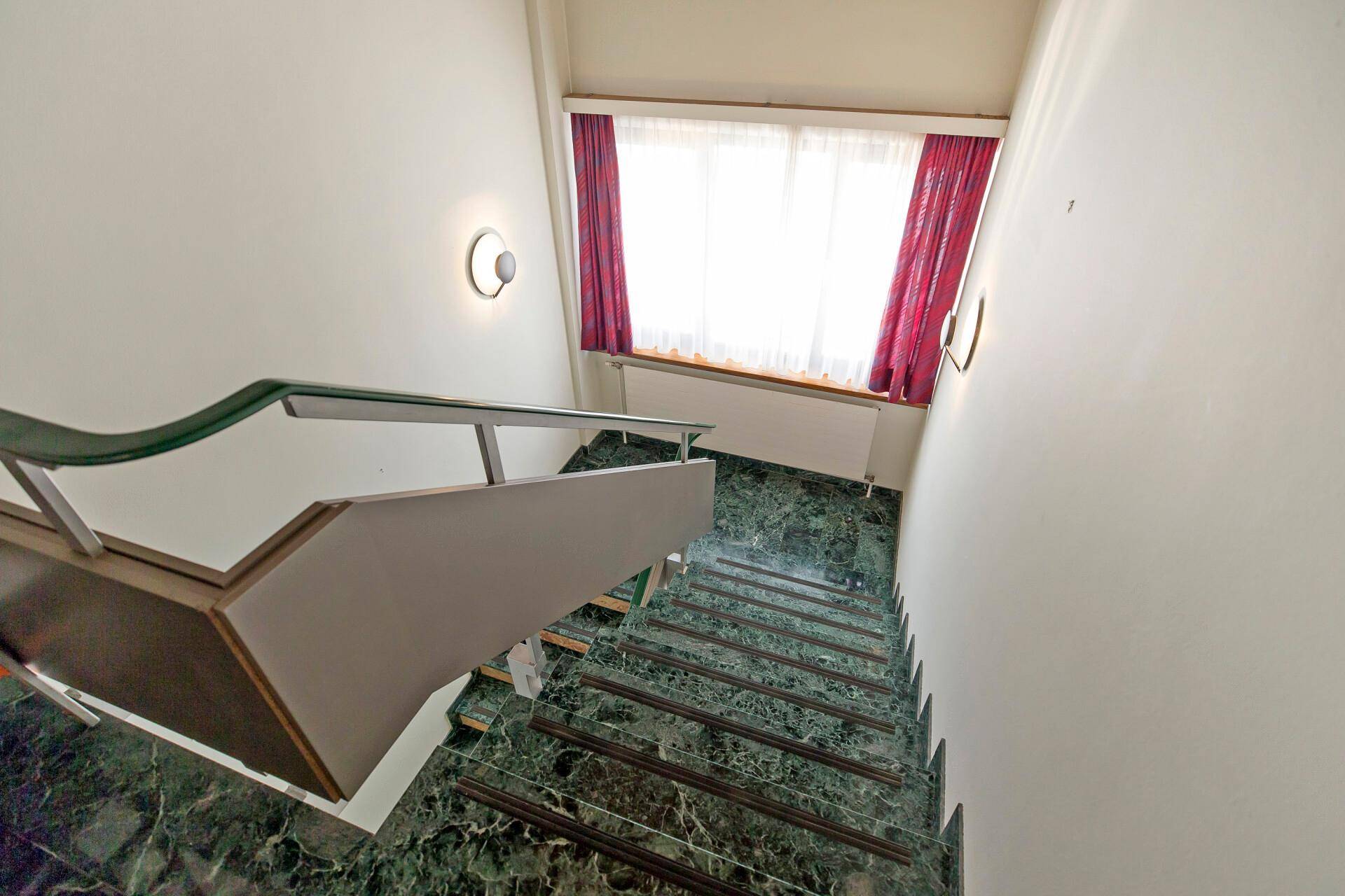 Treppenhaus mit elegantem Marmorboden, Glasgeländer und einem Fenster mit roten Vorhängen, das Licht hereinlässt.