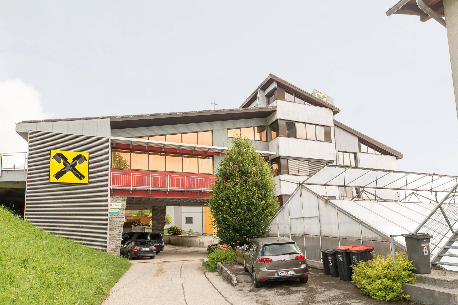 Außenansicht des Gebäudes mit moderner Architektur, Parkplätzen und angrenzenden Gewächshäusern in grüner Umgebung.