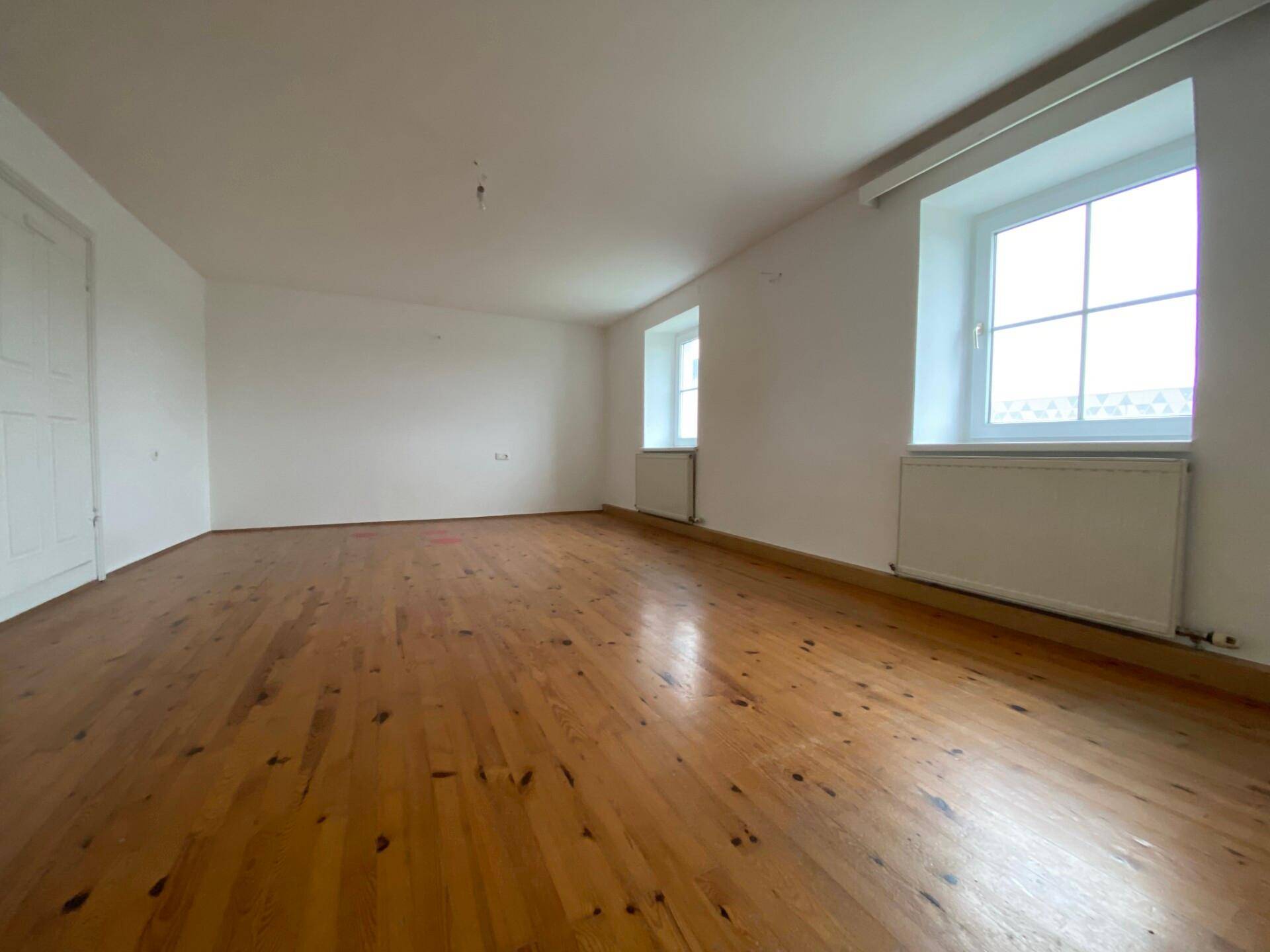 Heller, leerer Raum mit Holzdielenboden, weißen Wänden und zwei Fenstern, die viel Tageslicht hereinlassen.
