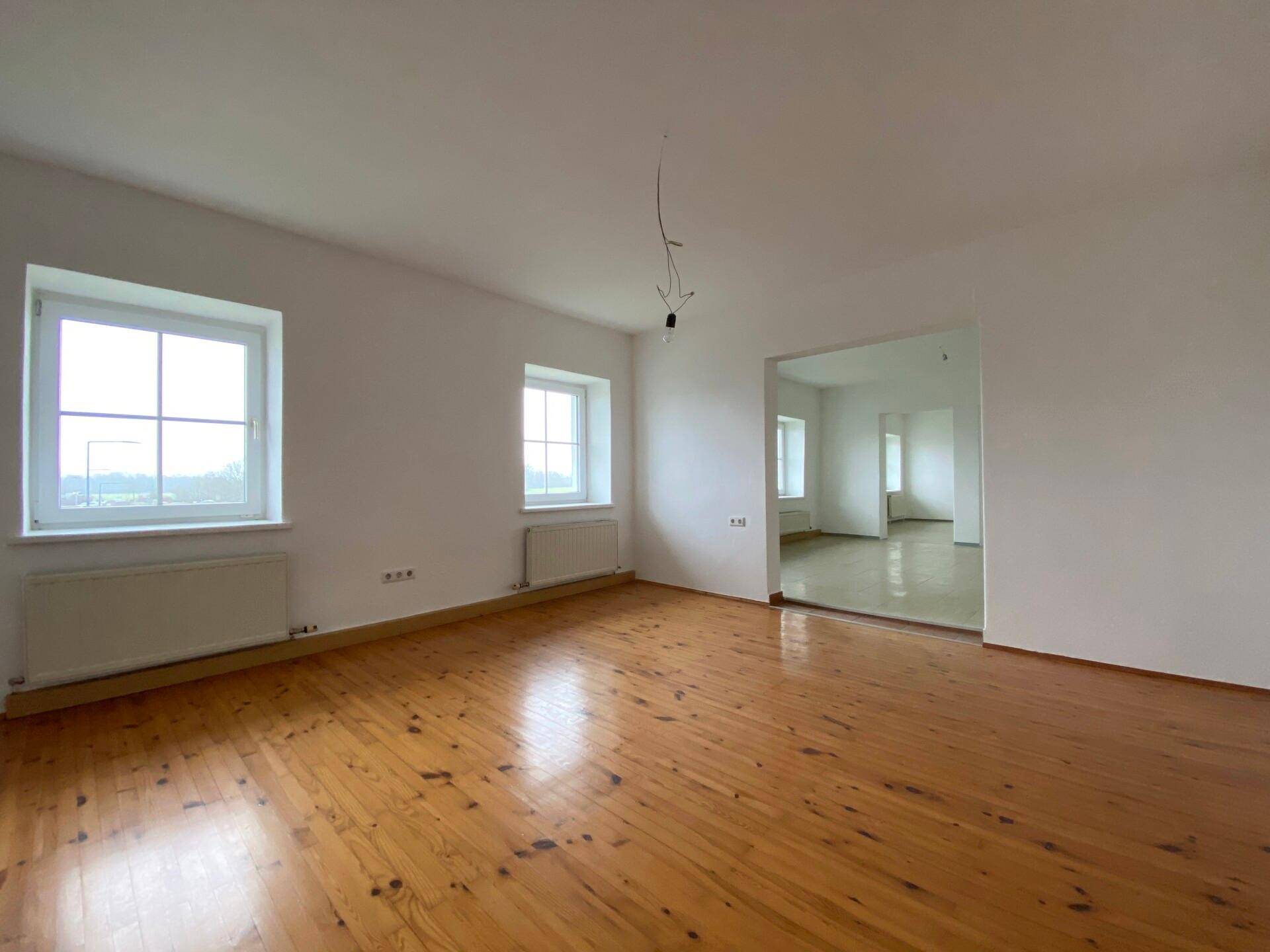 Heller, leerer Raum mit Holzdielenboden, weißen Wänden und einem offenen Durchgang zu einem weiteren Zimmer.