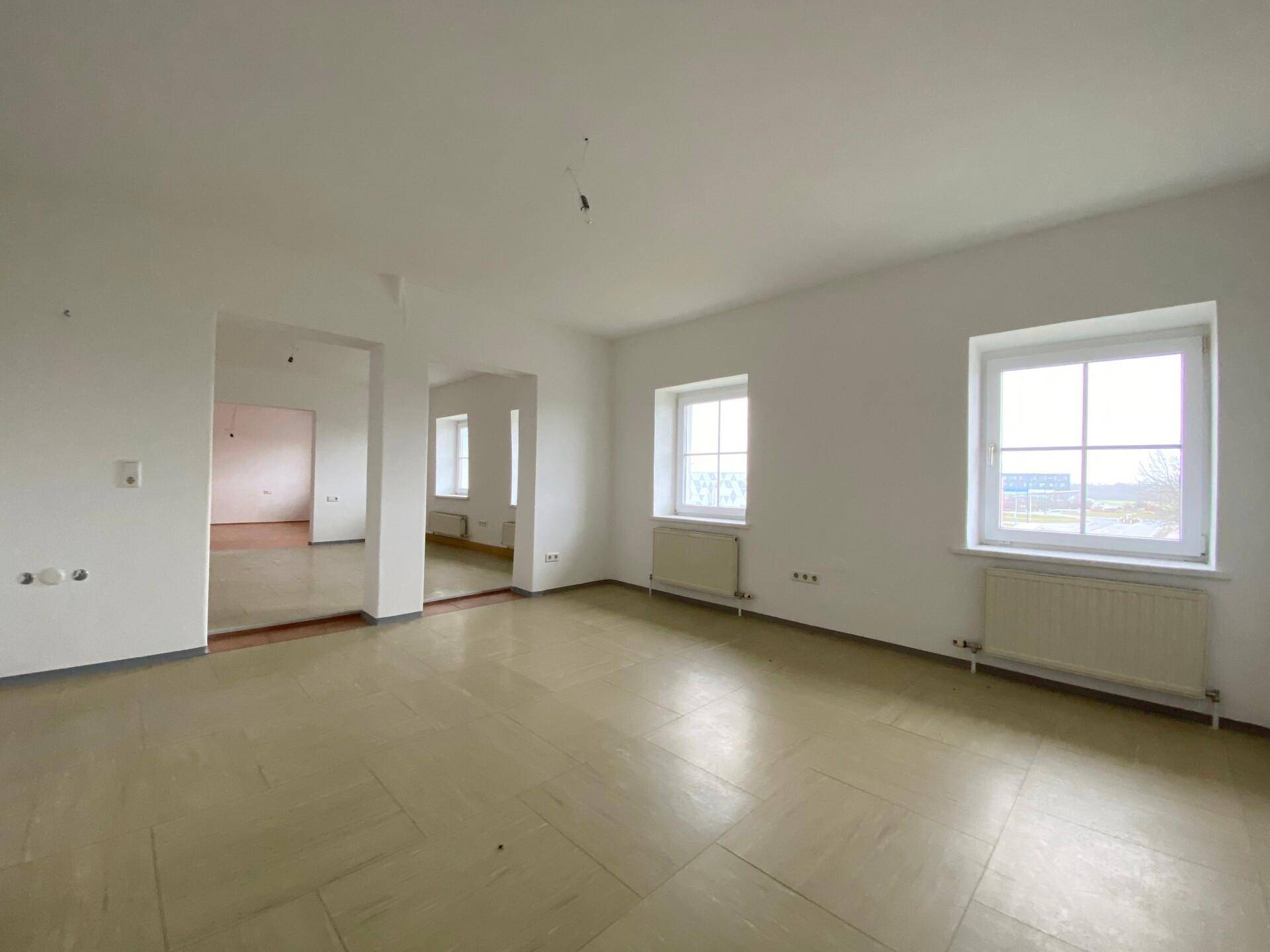 Heller, leerer Raum mit beigefarbenen Fliesen, weißen Wänden und einem offenen Durchgang zu einem weiteren Zimmer.