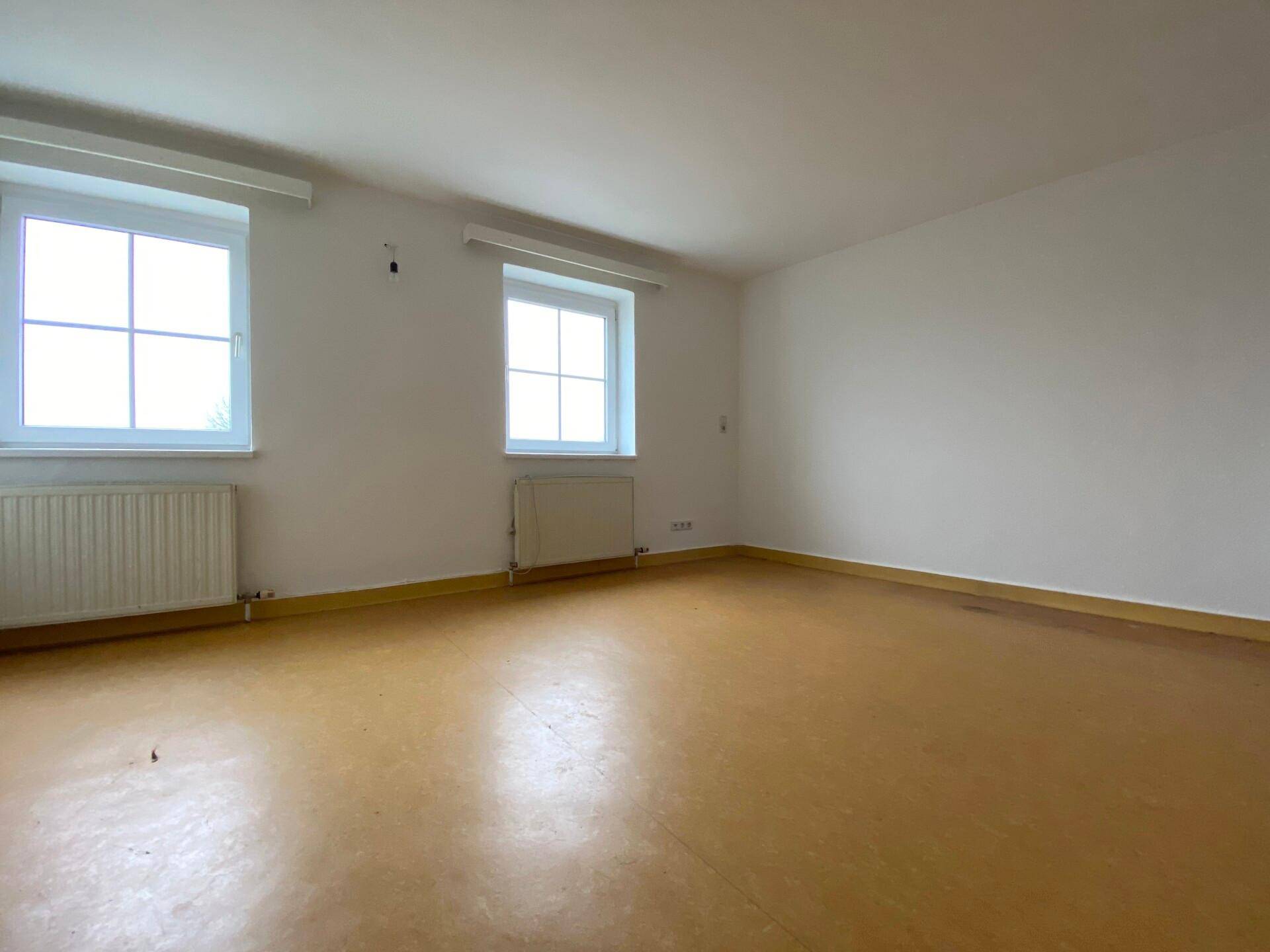 Geräumiger, leerer Raum mit braunem Boden, weißen Wänden und zwei Fenstern mit Heizkörpern darunter.