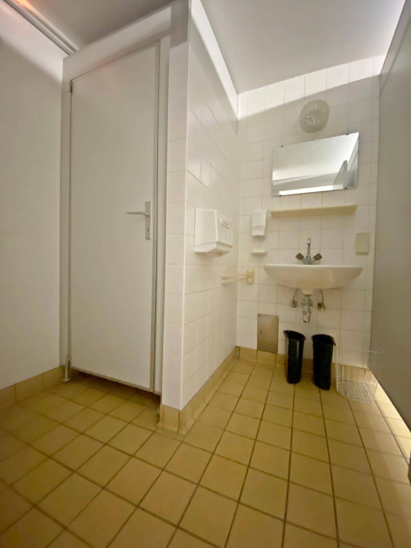Toilettenraum mit weißen Fliesenwänden, gelbbraunem Fliesenboden, einem Waschbecken und einer weißen Tür.
