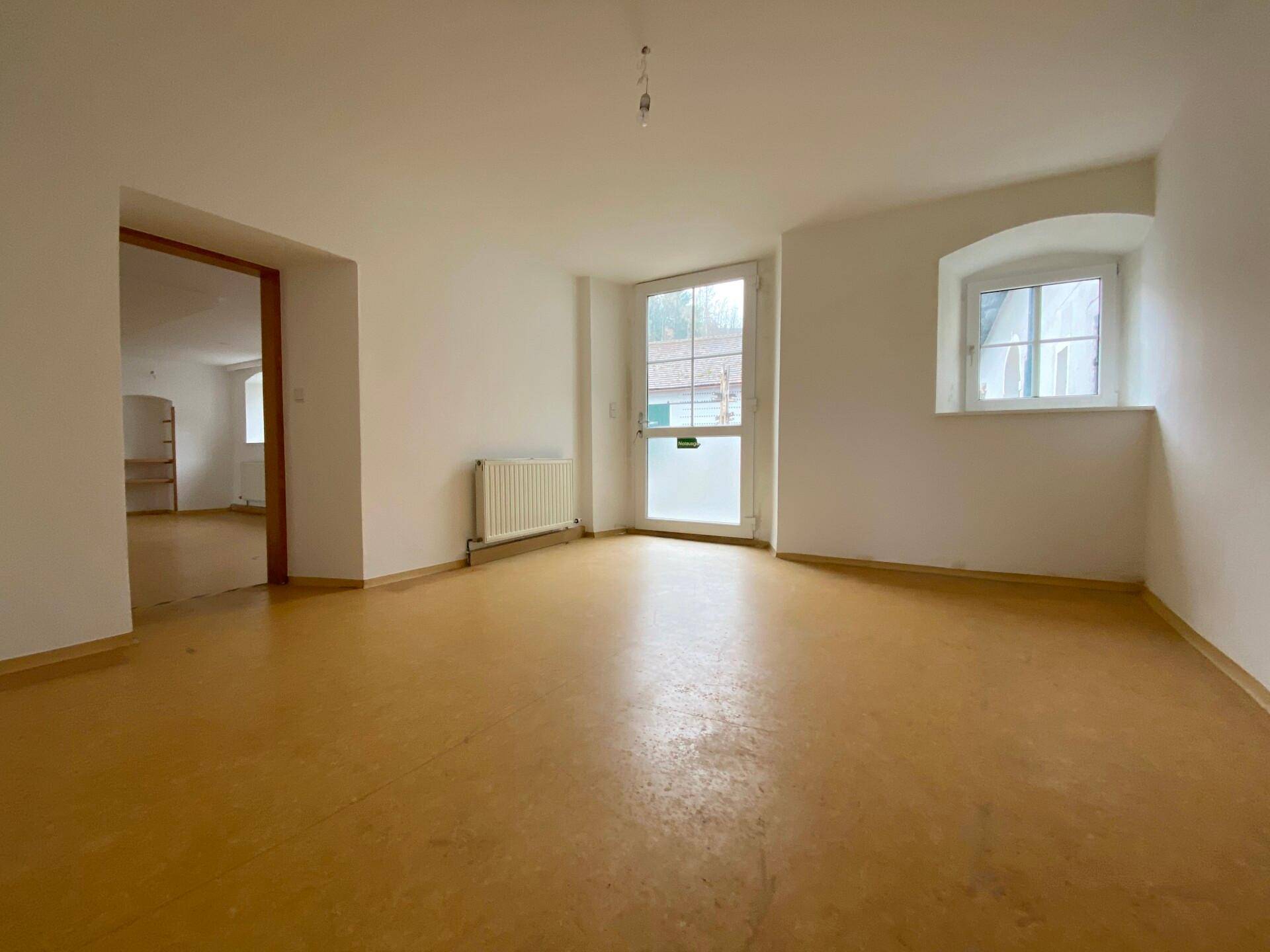 Heller, leerer Raum mit braunem Boden, weißen Wänden, einer Außentür und einem Rundbogenfenster.