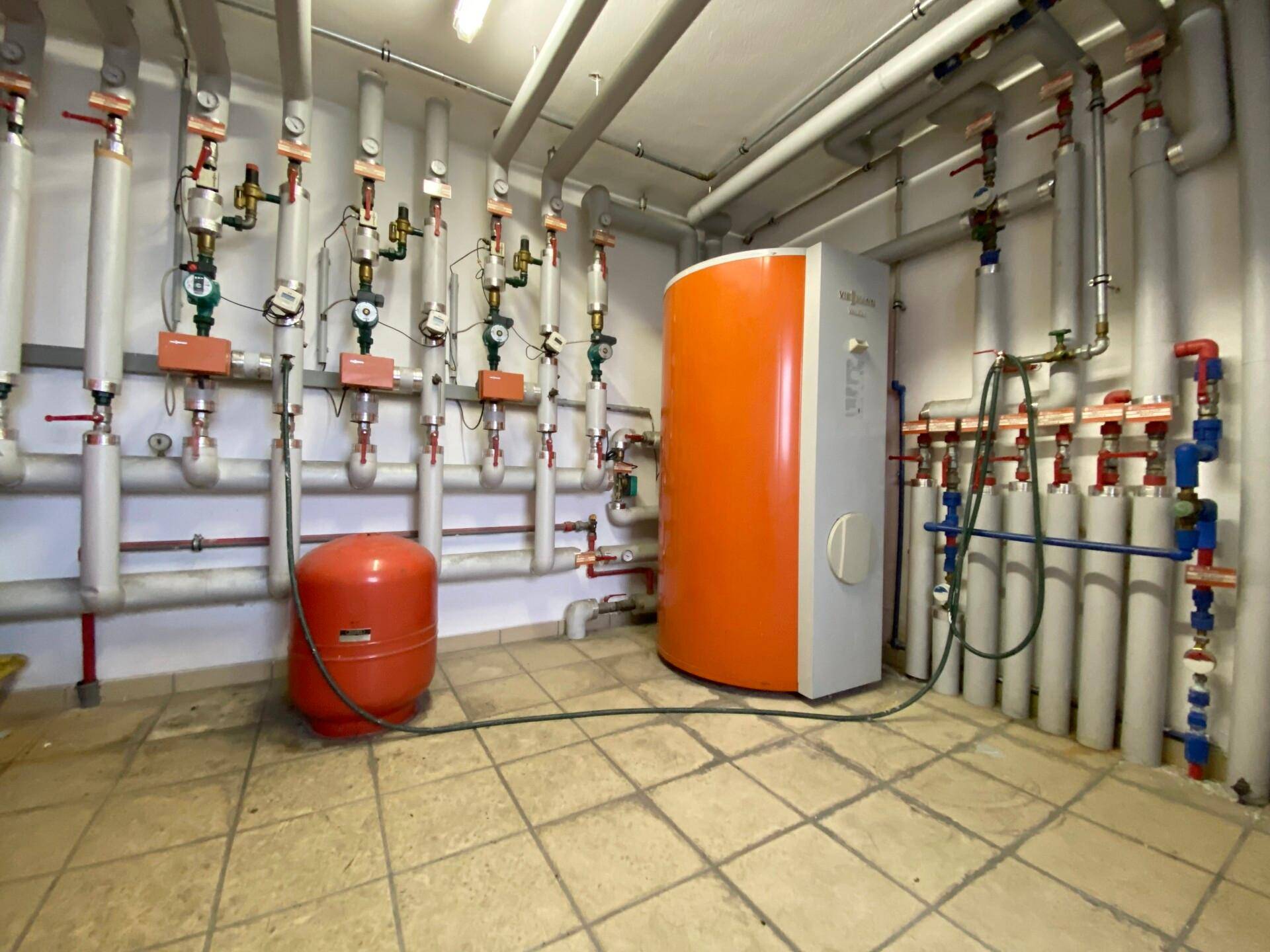 Heizungsraum mit einem großen orangefarbenen Warmwasserspeicher und einem komplexen Rohrsystem an den Wänden.