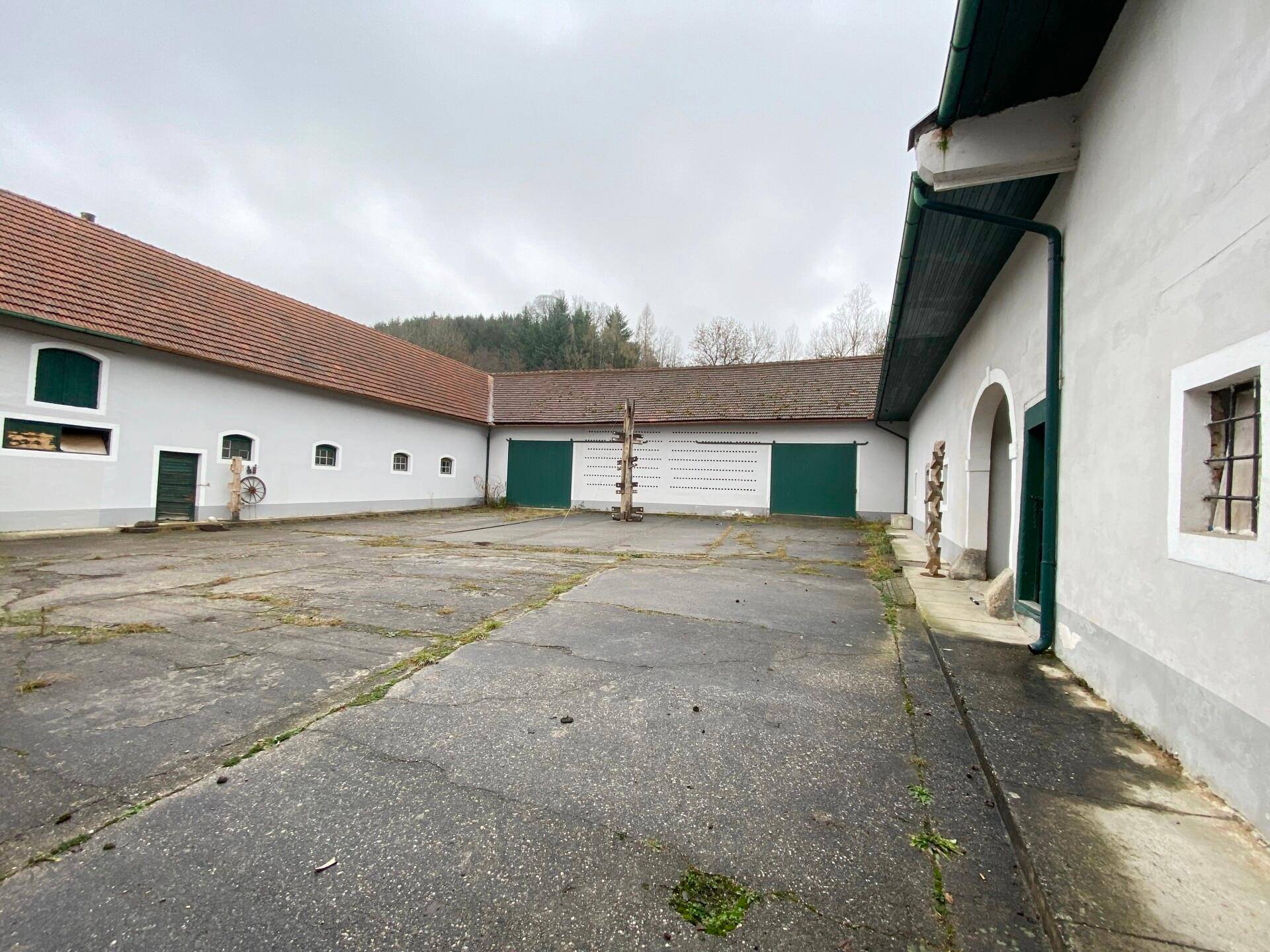 Weitläufiger Innenhof mit Gebäudefassaden, großen grünen Toren und einem Blick auf bewaldete Hügel.