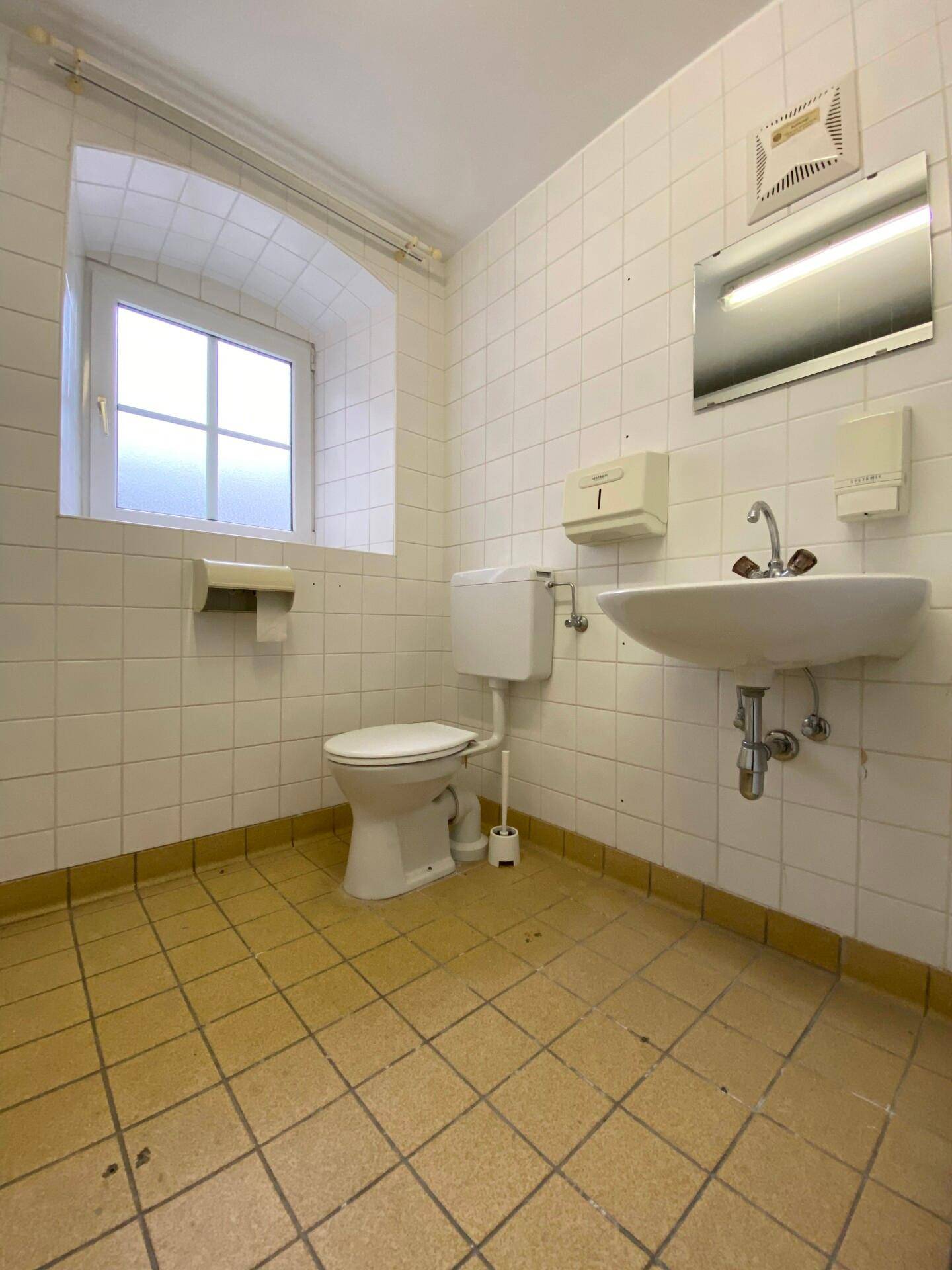 Toilettenraum mit weißen Fliesenwänden, gelbbraunem Fliesenboden, Toilette, Waschbecken und einem Fenster.