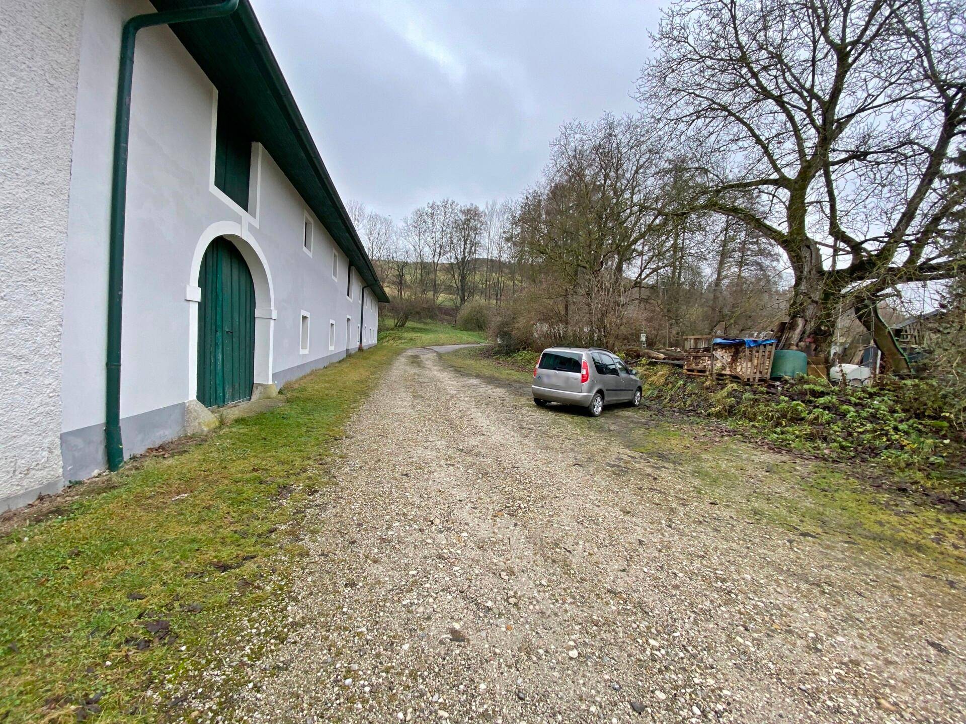 Seitenansicht des Gebäudes mit einem Schotterweg, einem geparkten Auto und alten Bäumen in ländlicher Umgebung.