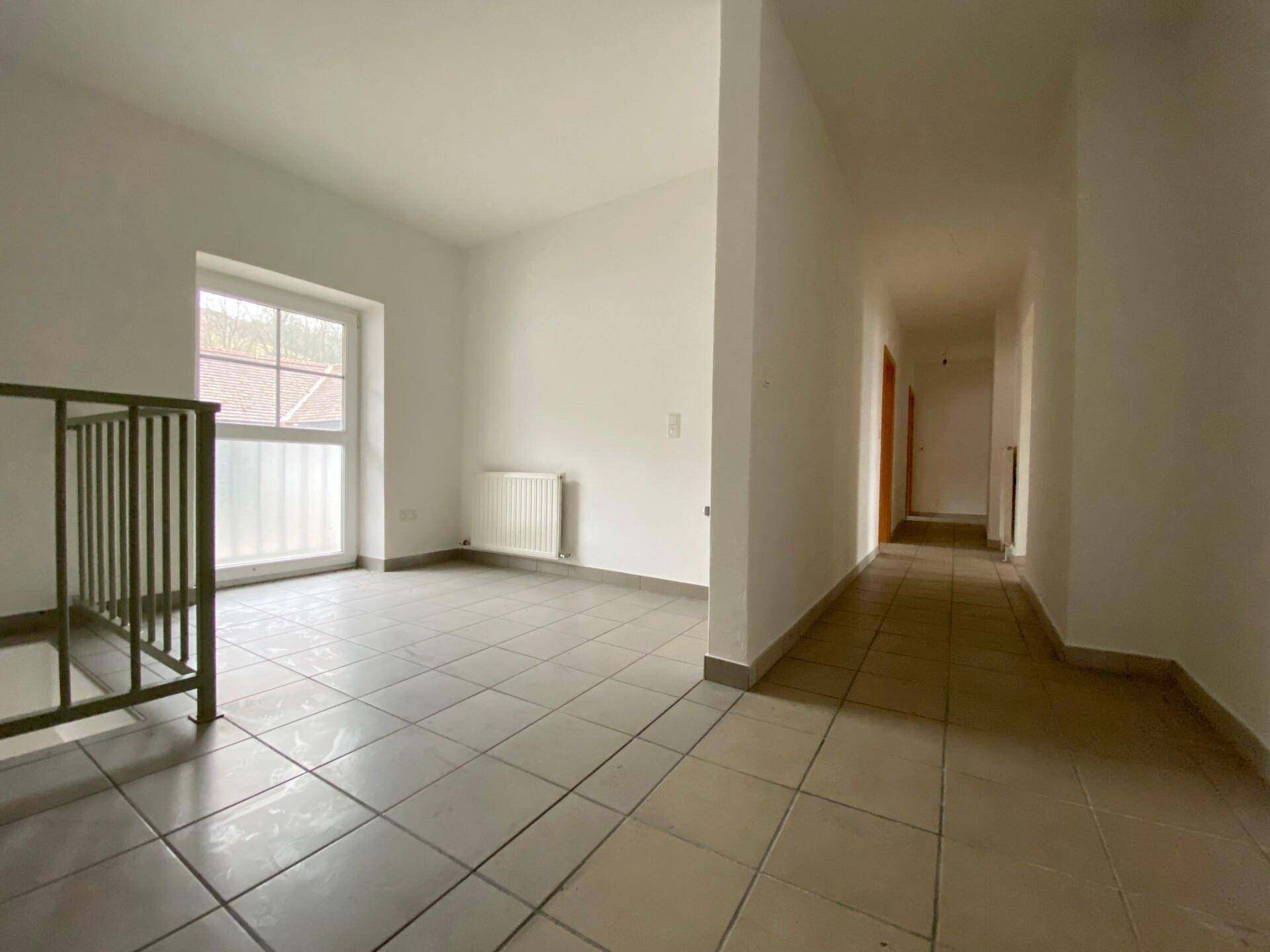 Heller Flur mit beigefarbenen Fliesen, weißen Wänden, einem Fenster und einem Metallgeländer für die Treppe.