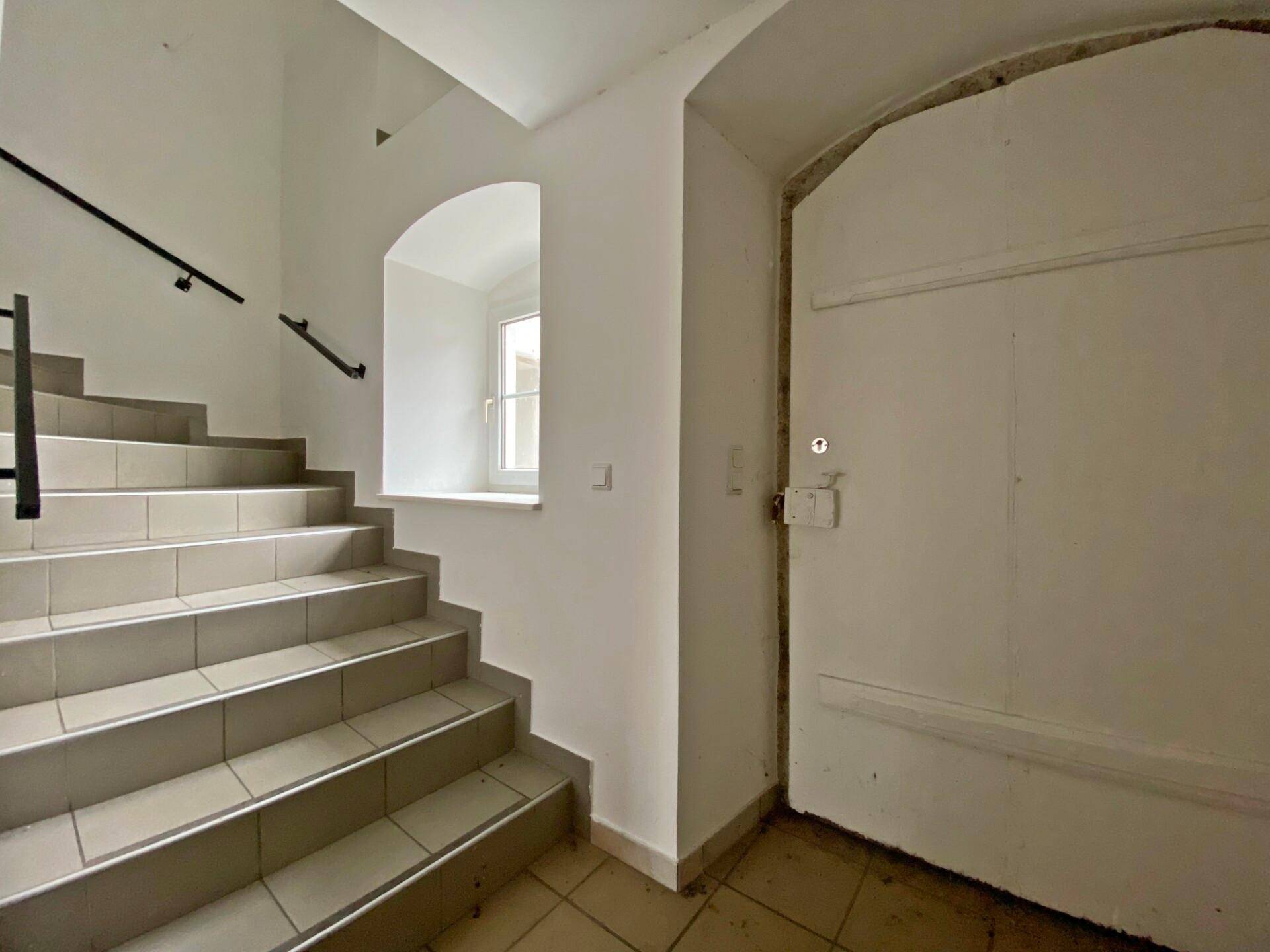 Treppenhaus mit beigefarbenen Fliesenstufen, weißen Wänden, einer großen Tür und einem Rundbogenfenster.