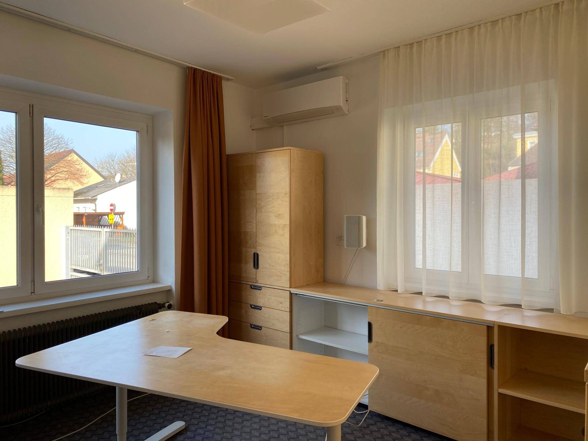 Helles Bürozimmer mit zwei Fenstern, Holzmöbeln und einer Klimaanlage.