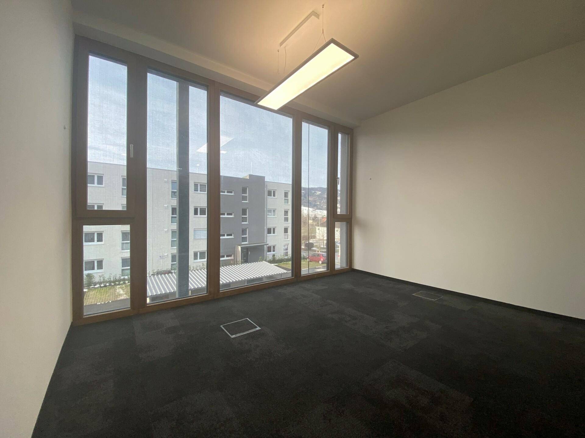 Heller Büroraum mit großen Fenstern, die viel Tageslicht hereinlassen und einen Blick auf umliegende Gebäude bieten.