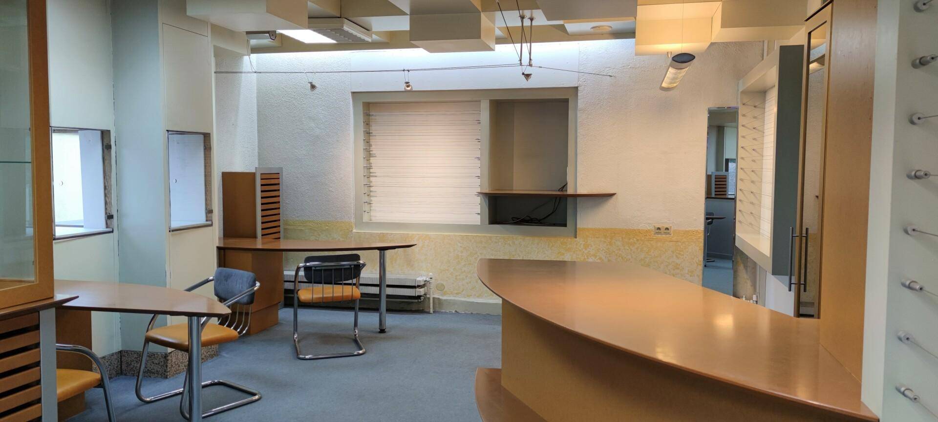 Büro- oder Empfangsbereich mit geschwungener Holztheke, Schreibtischen und Fenstern mit Jalousien.
