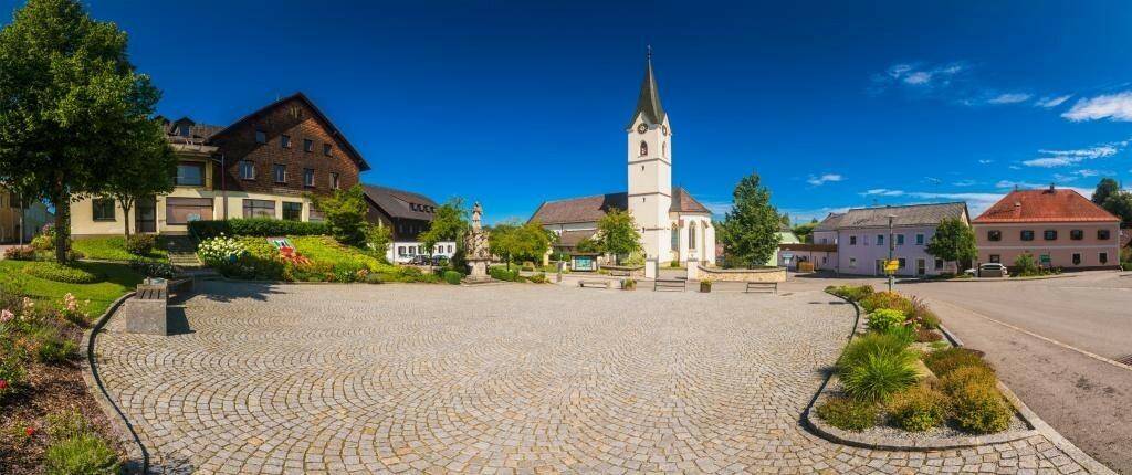Panoramablick auf einen gepflasterten Dorfplatz mit einer Kirche und traditionellen Gebäuden unter blauem Himmel.