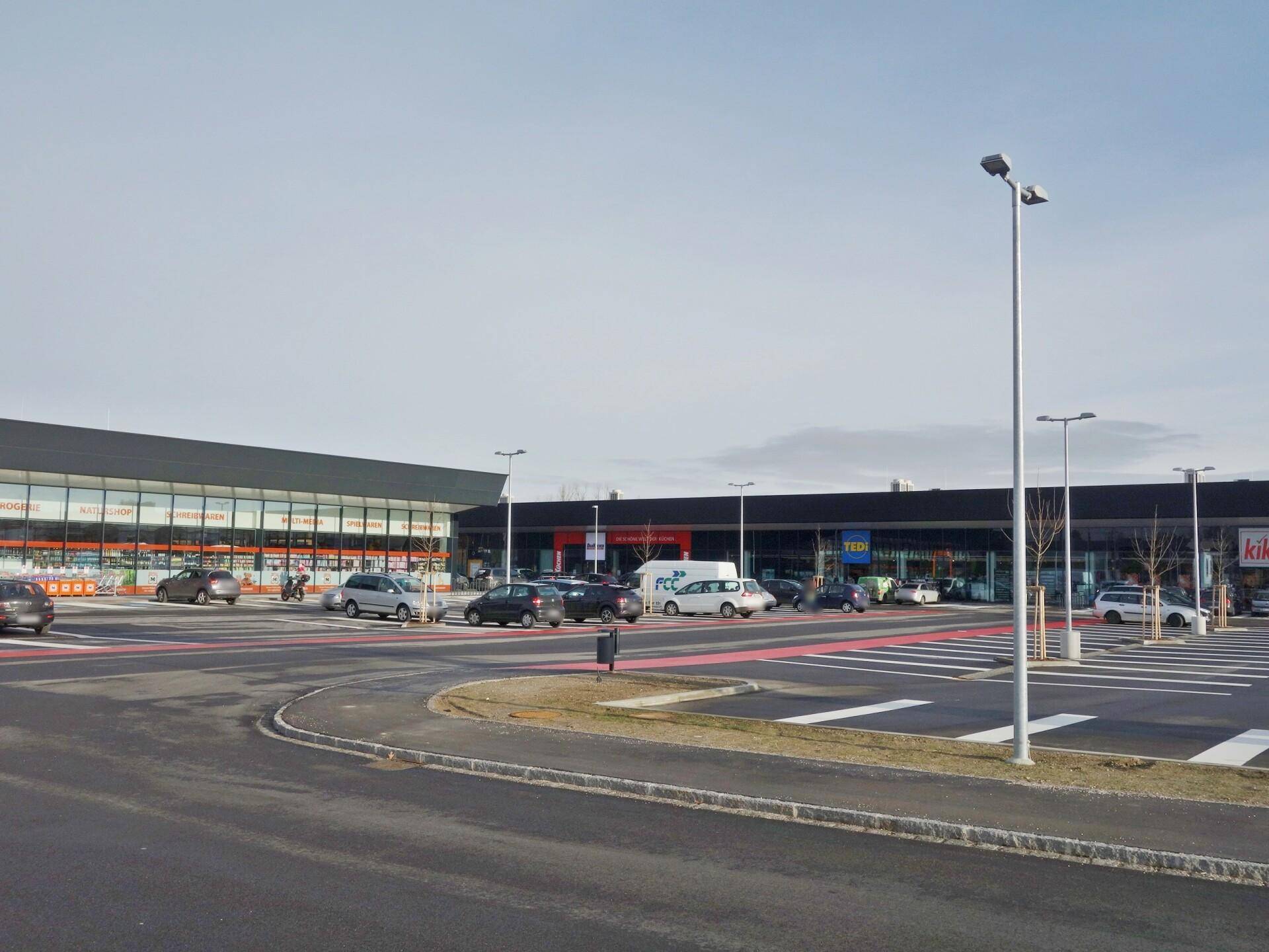 Weitläufiger Parkplatz vor einem modernen Einkaufszentrum mit verschiedenen Geschäften und klarem Himmel.