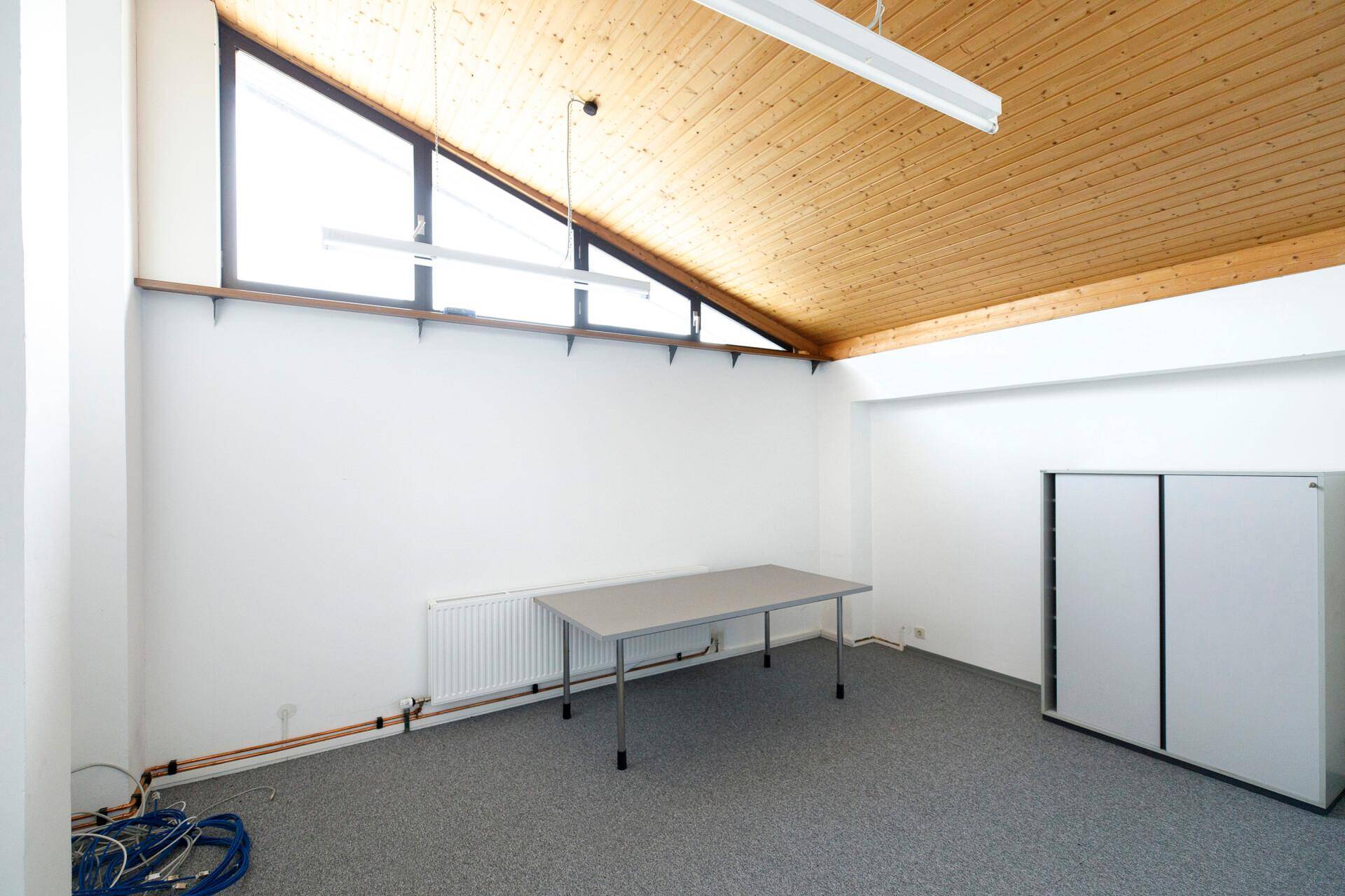 Kleinerer Raum mit Holzbalkendecke, schrägen Fenstern und grauem Teppichboden, ideal als Einzelbüro.