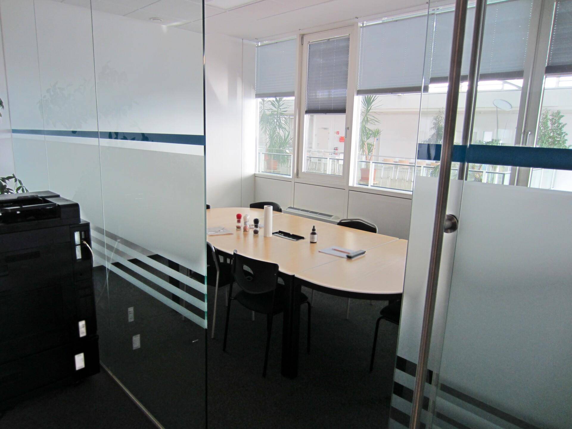 Besprechungsraum mit ovalem Tisch, Stühlen und Fensterfront mit Jalousien, durch Glastür sichtbar.