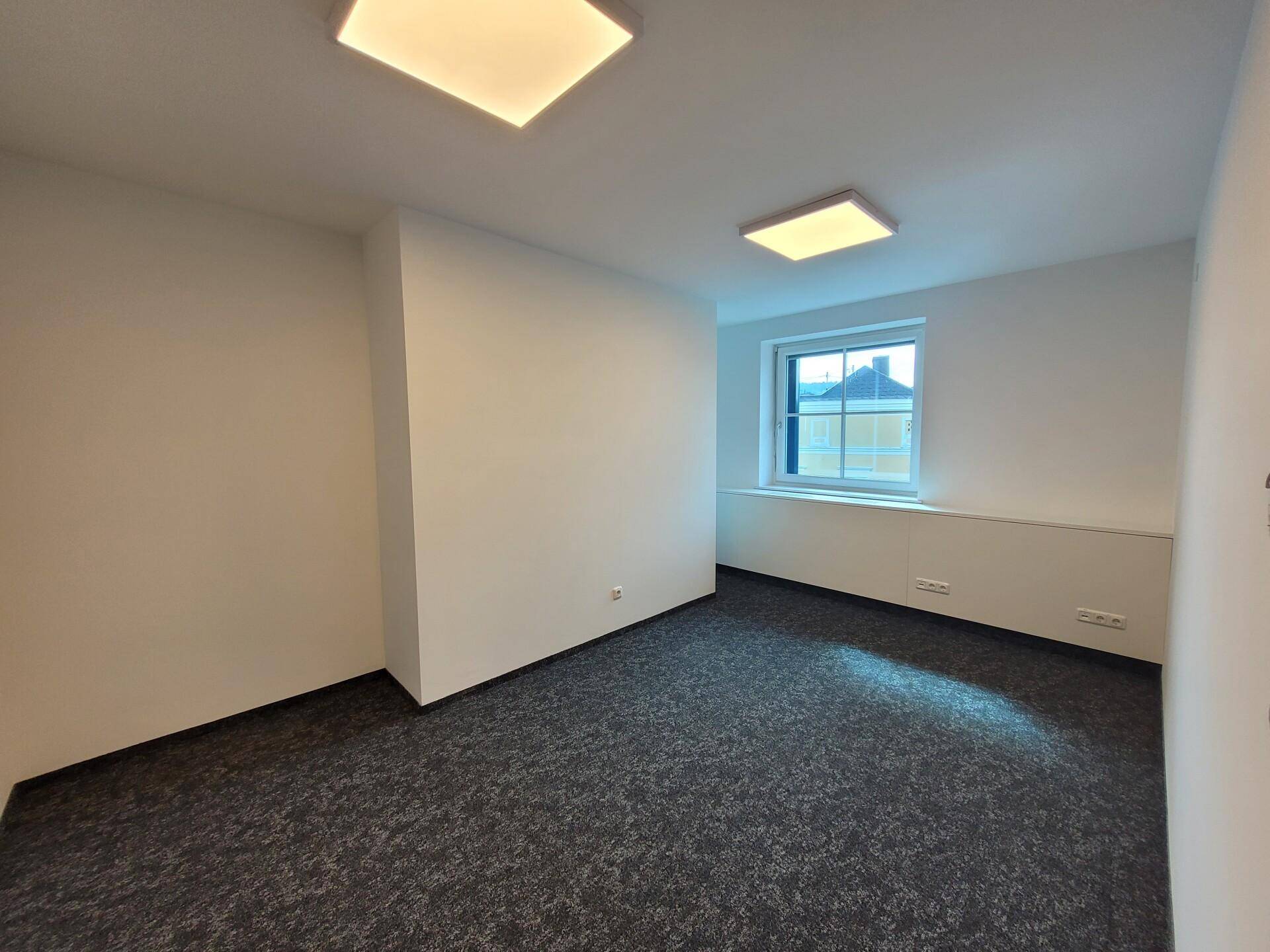Leerer Büroraum mit Teppichboden und einem Fenster, das Tageslicht hereinlässt.