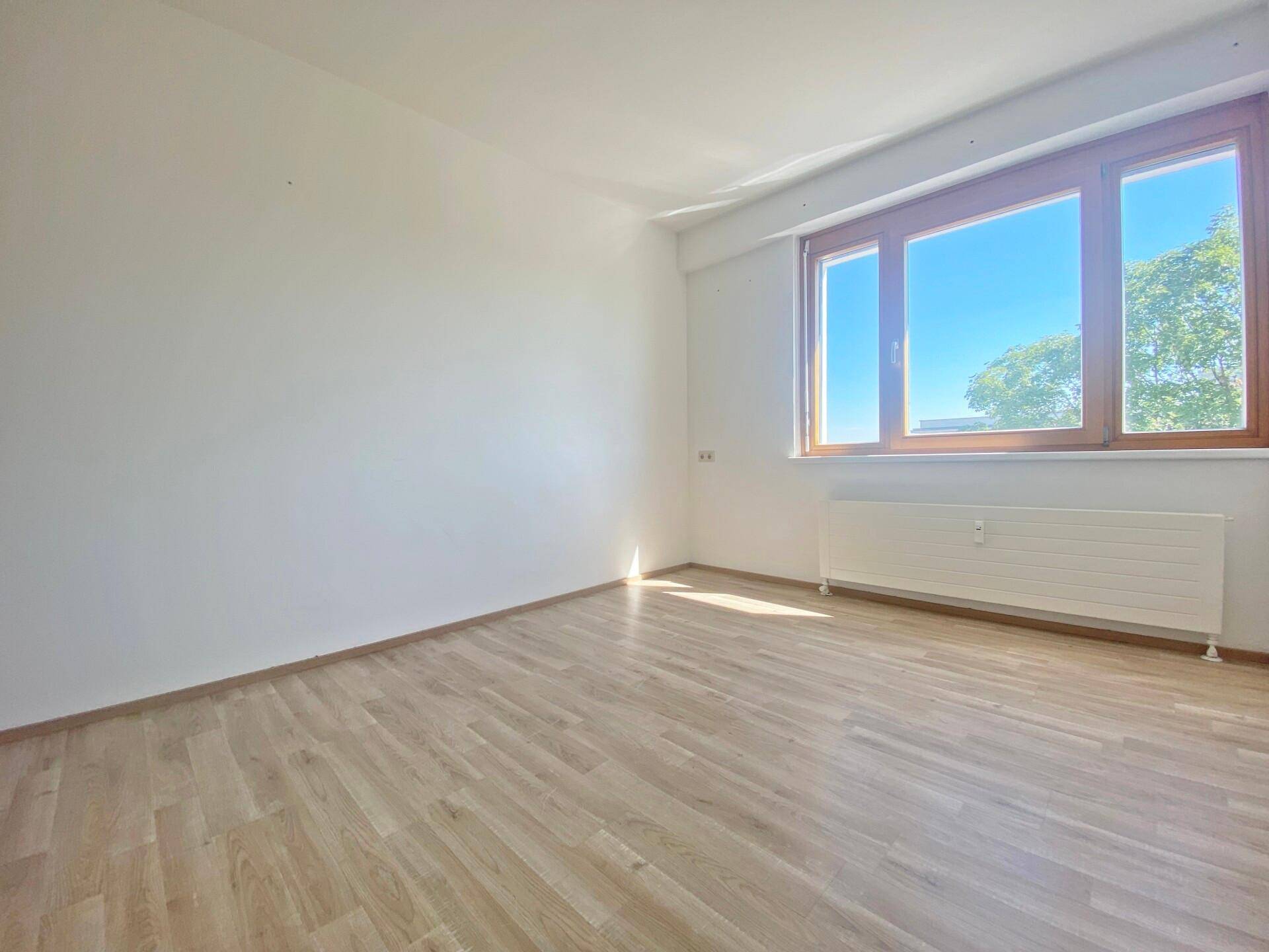 Leeres, helles Zimmer mit Laminatboden und großem Fenster, bereit zur individuellen Gestaltung.