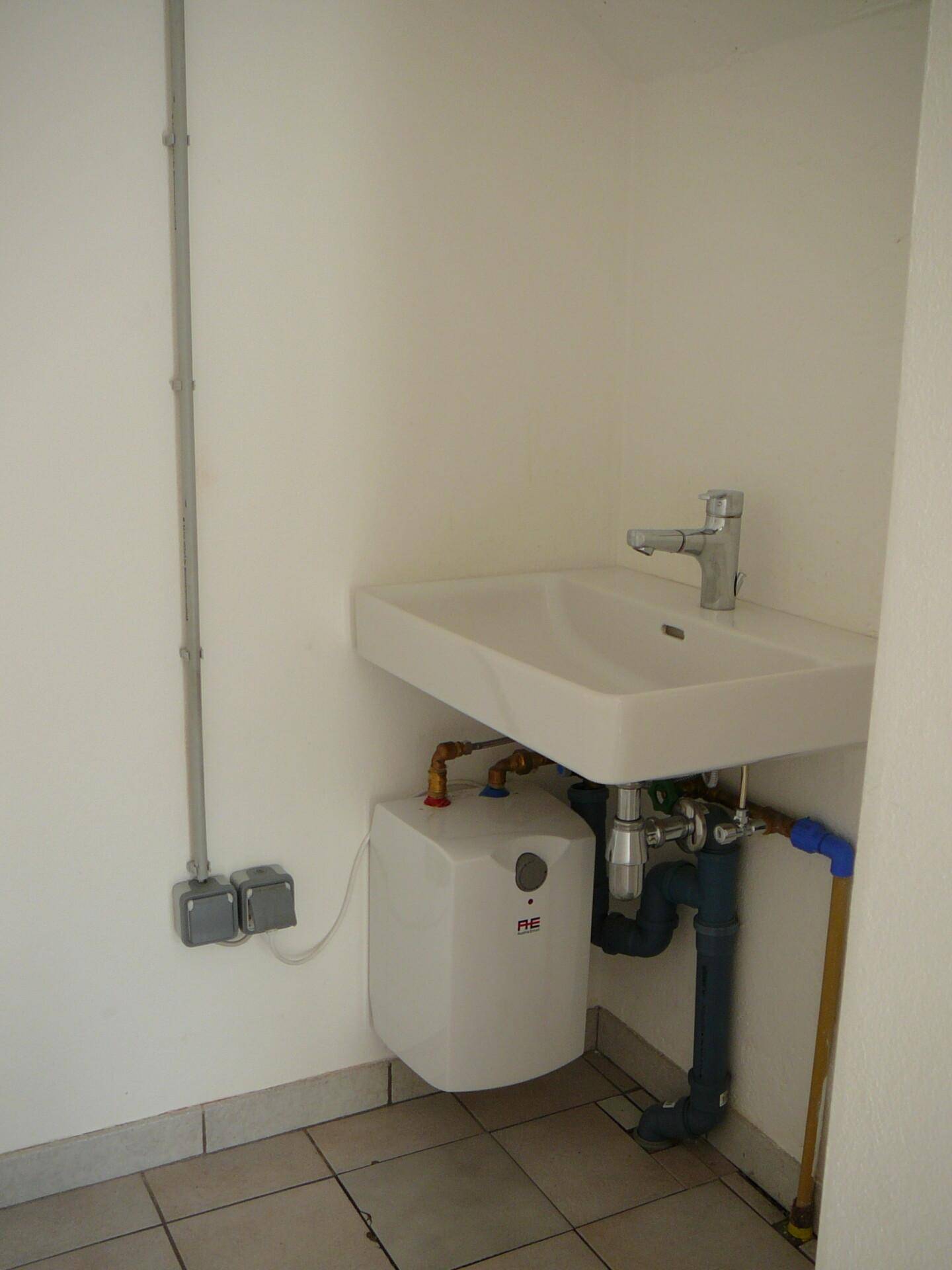 Einfaches Badezimmer mit Waschbecken, Untertischboiler und sichtbaren Rohrleitungen an der Wand.