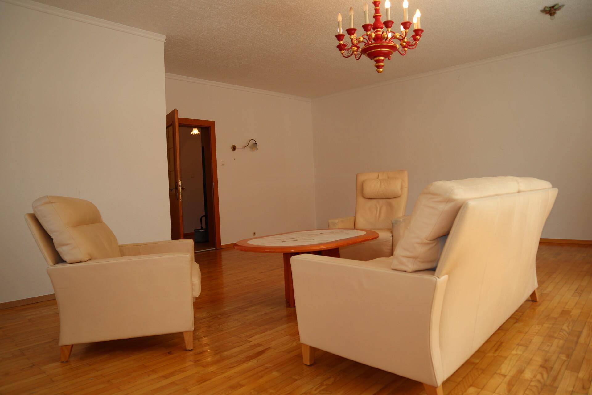 Geräumiges Wohnzimmer mit hellem Holzfußboden, beigen Polstermöbeln und einem roten Kronleuchter.