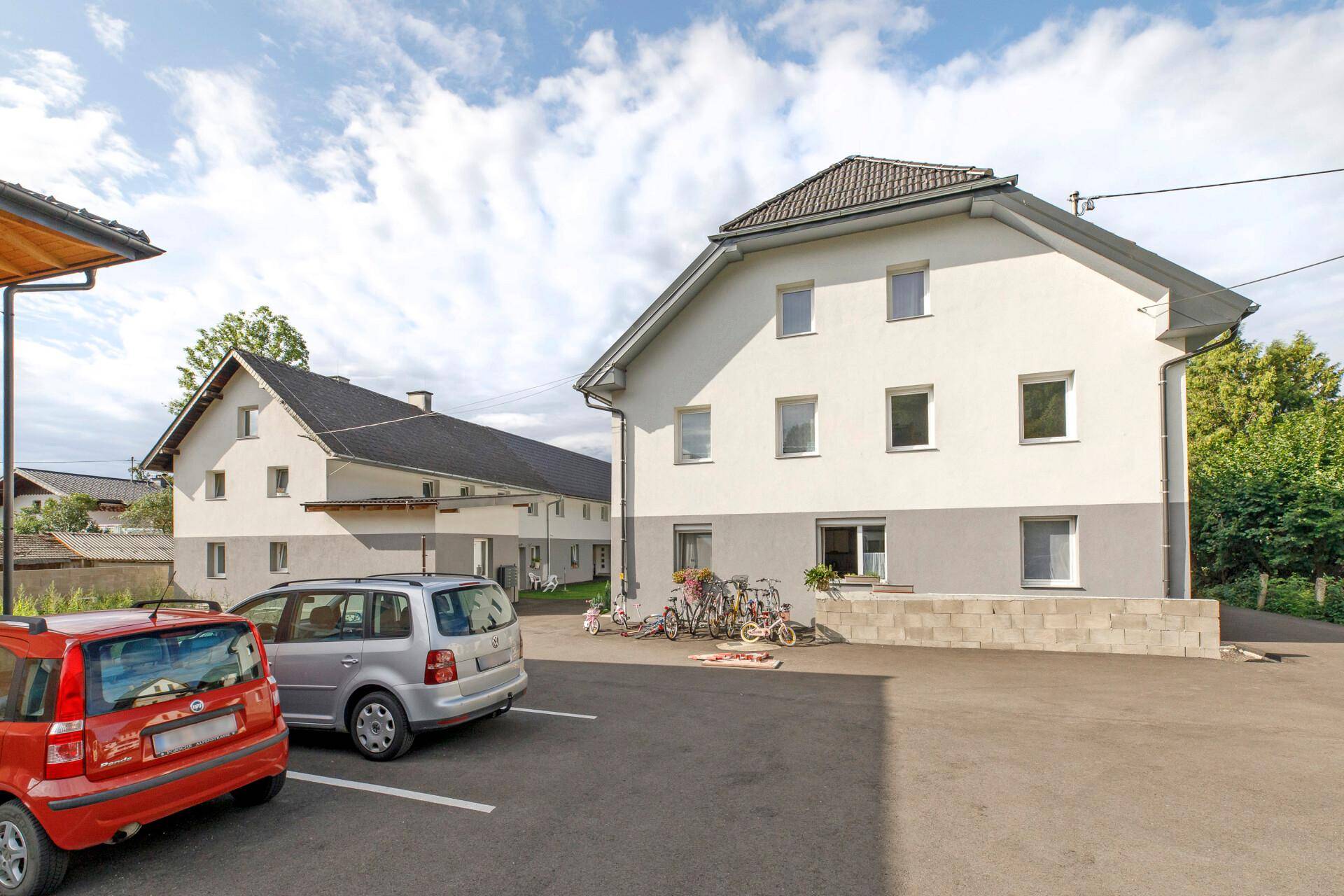 Zwei moderne Mehrfamilienhäuser mit Satteldach und einem großen Parkplatz davor unter blauem Himmel.