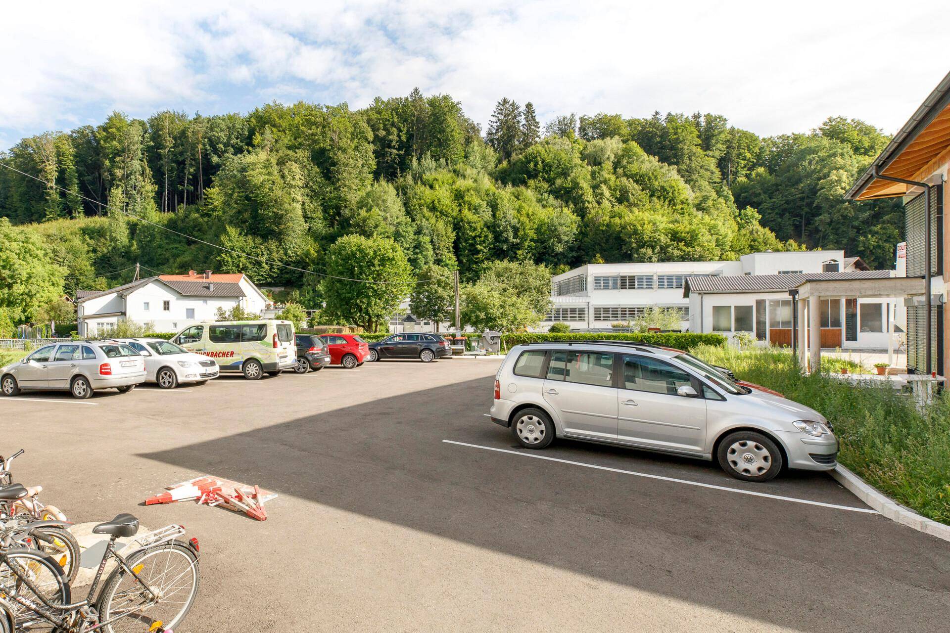 Großer Parkplatz vor dem Gebäude mit Blick auf einen bewaldeten Hügel und weitere Häuser in der Umgebung.