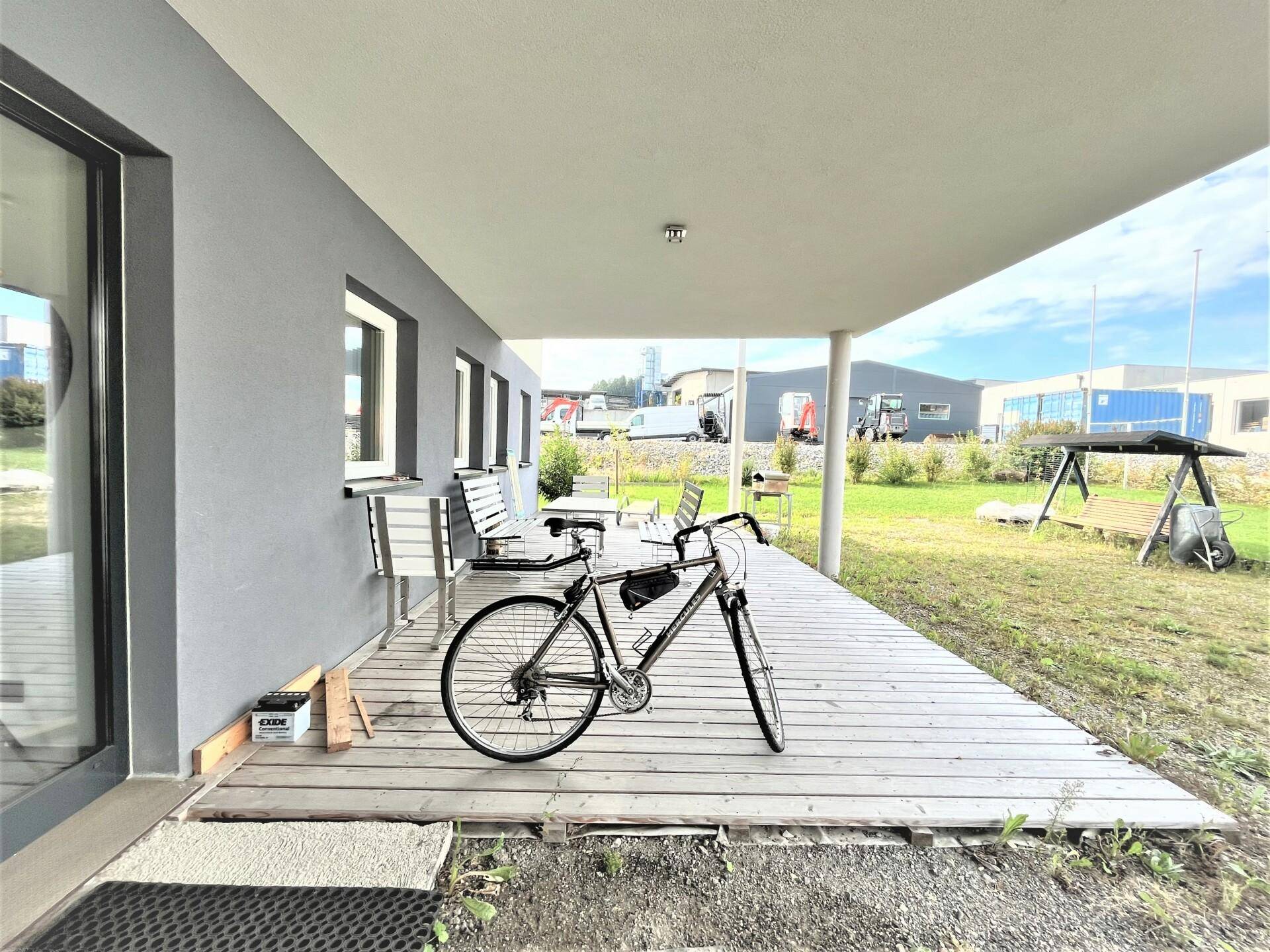 Überdachte Holzterrasse mit Sitzgelegenheiten und einem Fahrrad, umgeben von einer Rasenfläche.