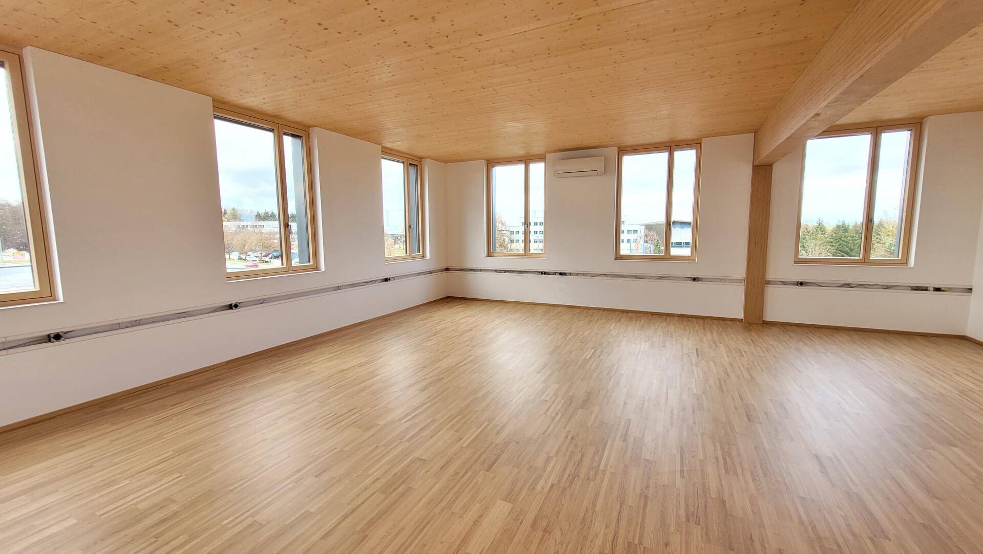 Heller, leerer Mehrzweckraum mit Holzboden, Holzdecke, vielen Fenstern und Klimaanlage.