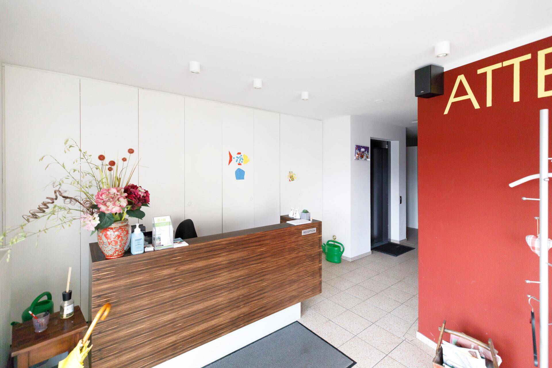 Empfangsbereich mit Holztheke, weißen Wänden und einem Aufzug im Hintergrund.