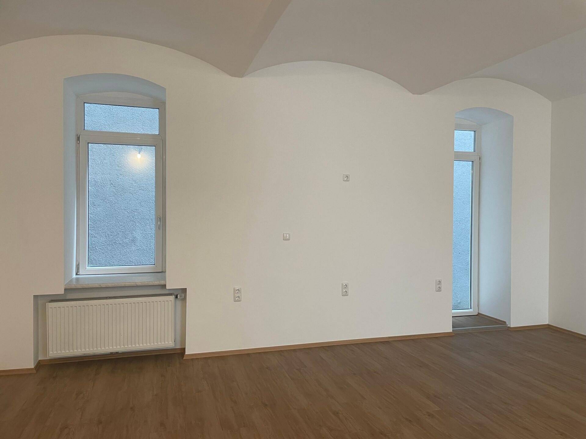 Leerer Raum mit gewölbter Decke, zwei Fenstern und modernem Holzboden.