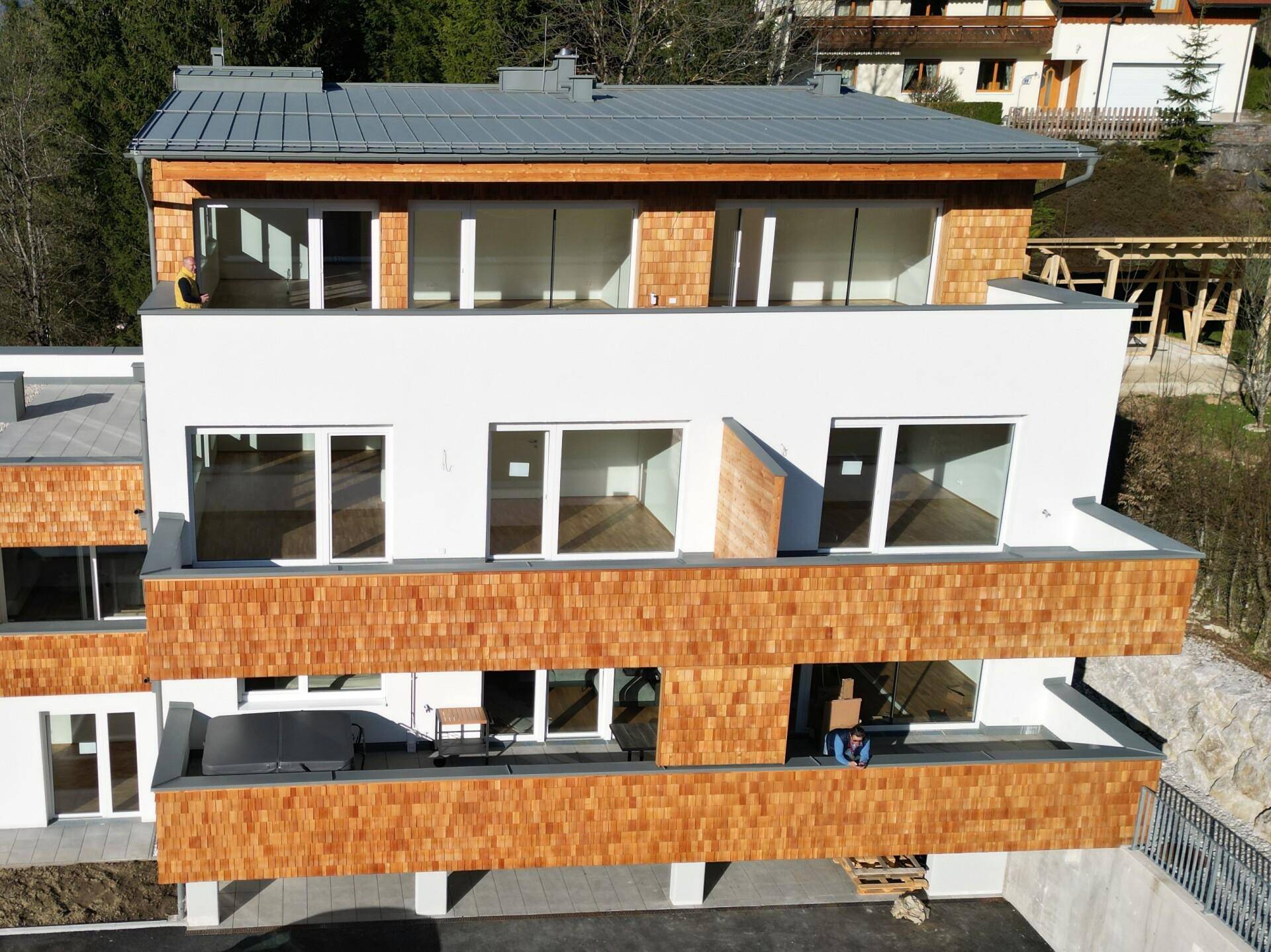 Modernes Gebäude mit Holzfassadenelementen und mehreren Balkonen in alpiner Umgebung, umgeben von Natur.