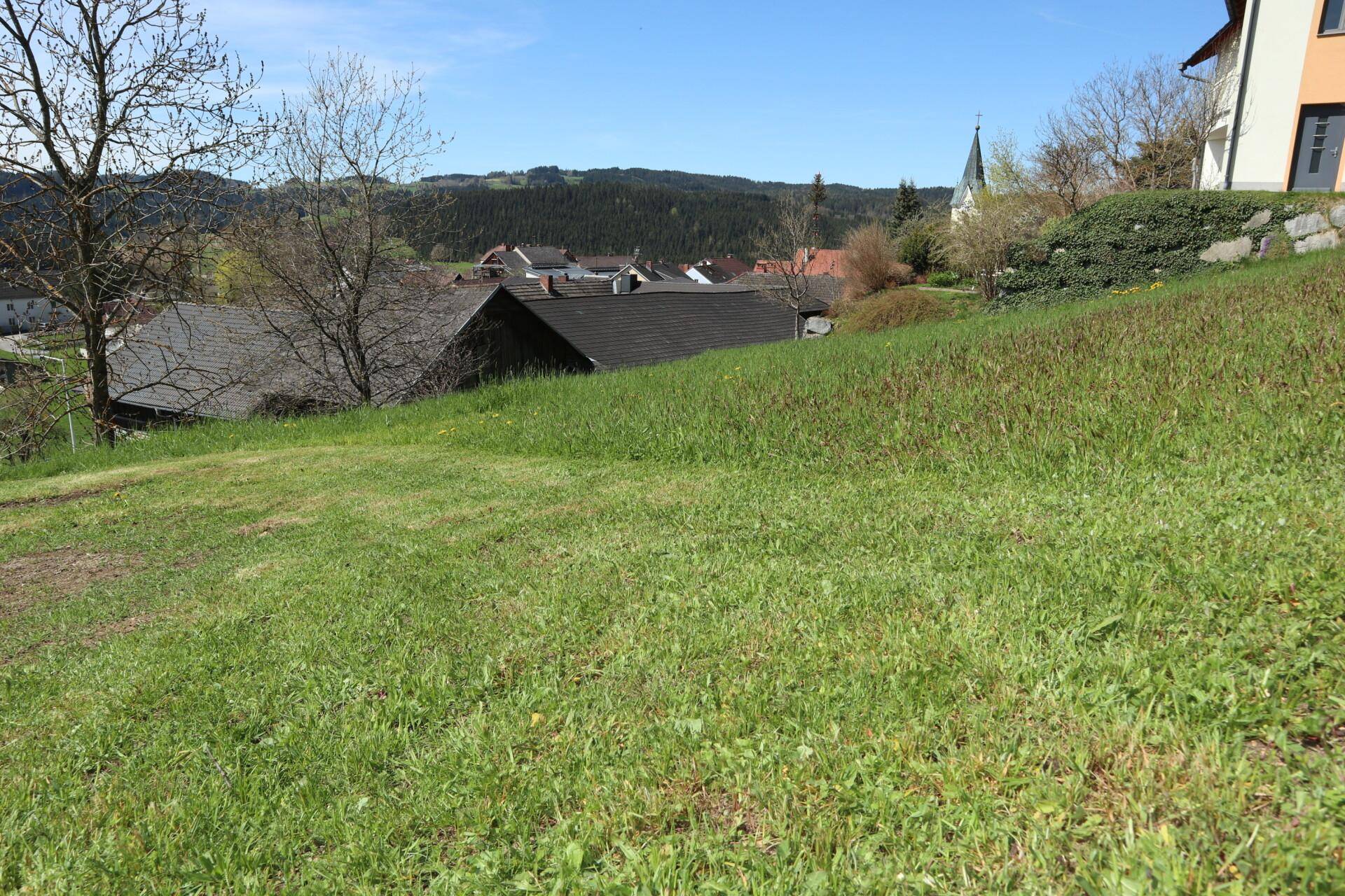 Panoramablick über eine grüne Wiese auf ein malerisches Dorf mit Kirche und bewaldeten Hügeln.