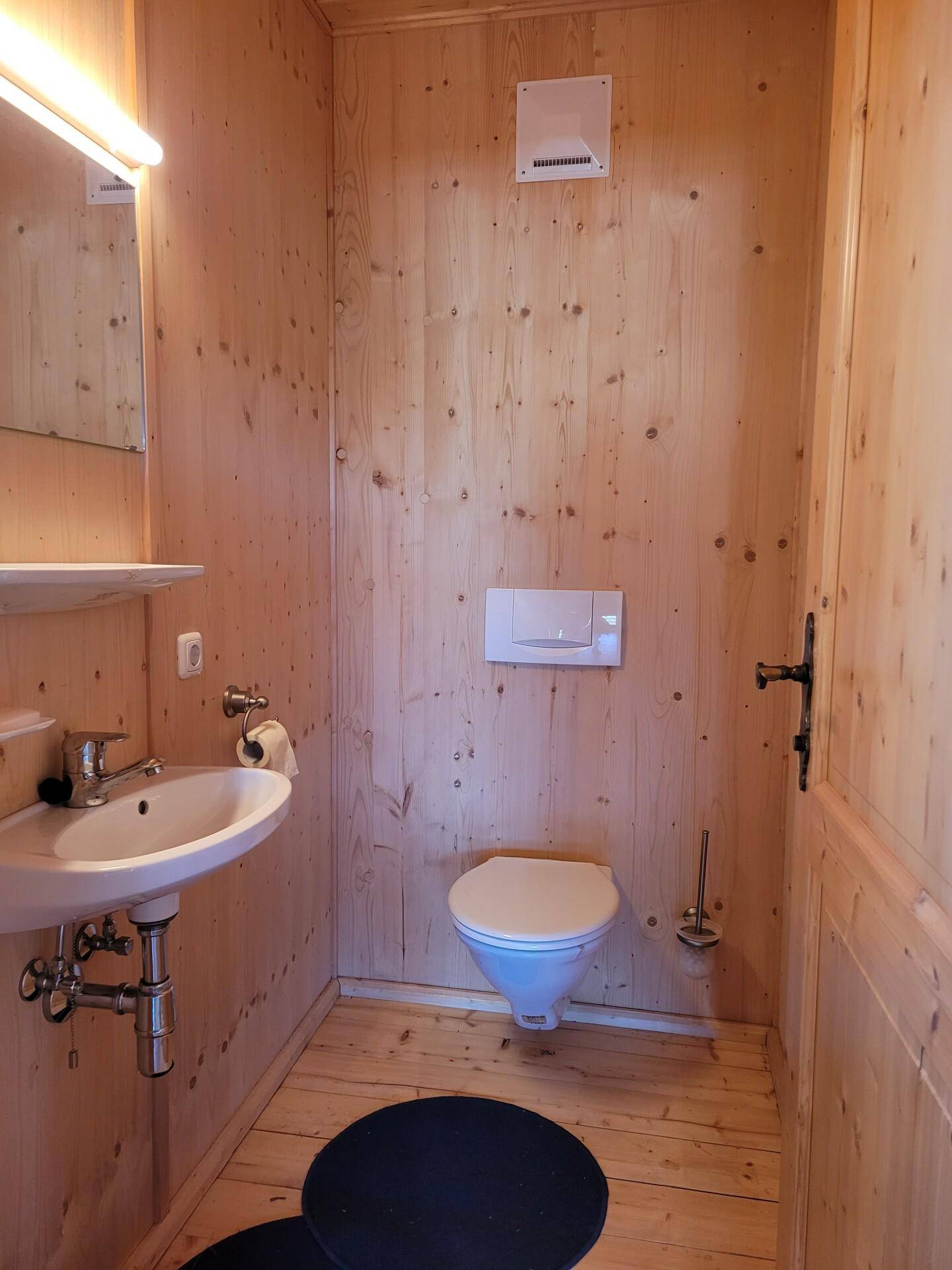 Kleine Gästetoilette mit Holzwänden, wandhängendem WC, Waschbecken und einem Abluftventilator.