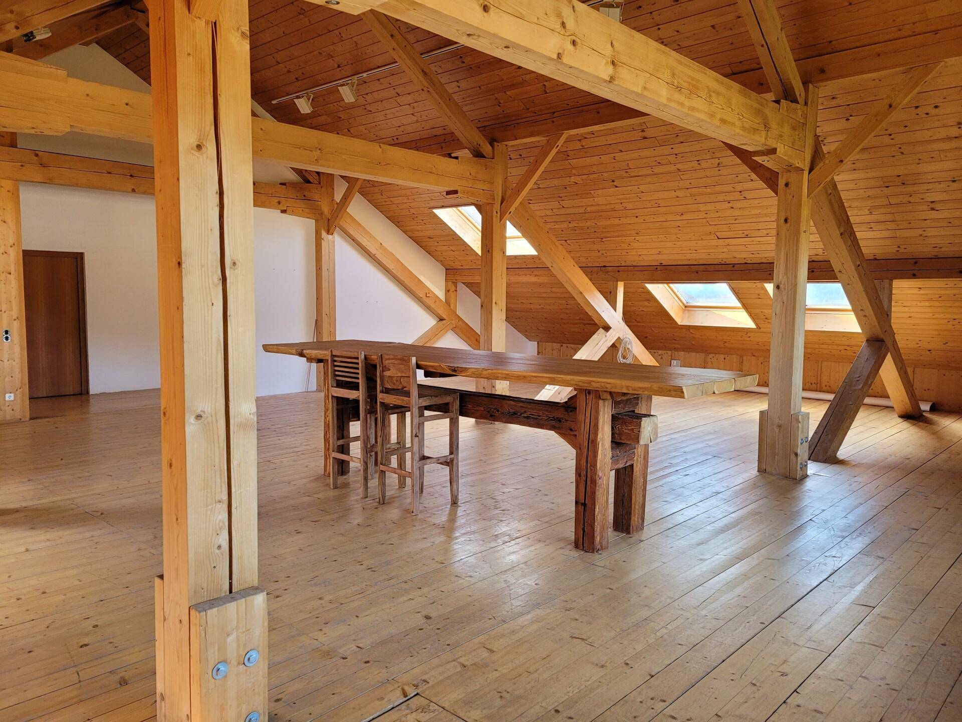 Weitläufiger offener Raum mit beeindruckender Holzkonstruktion, Holzboden und einem massiven Holztisch.