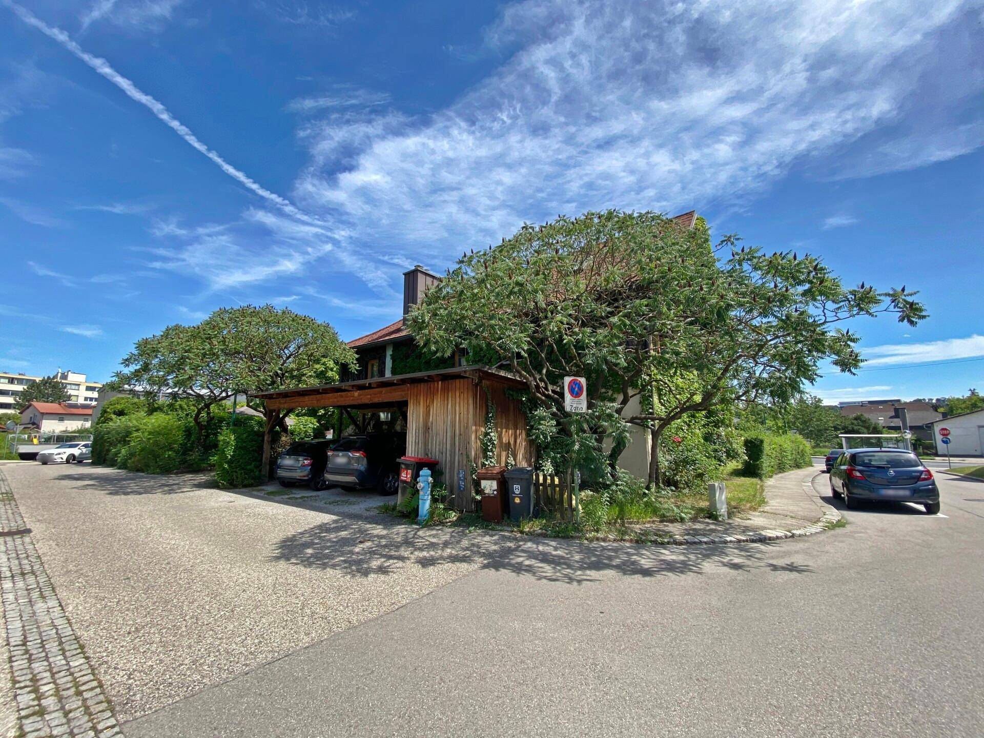 Gebäudeansicht von der Straße mit einem überdachten Carport und viel Grün.