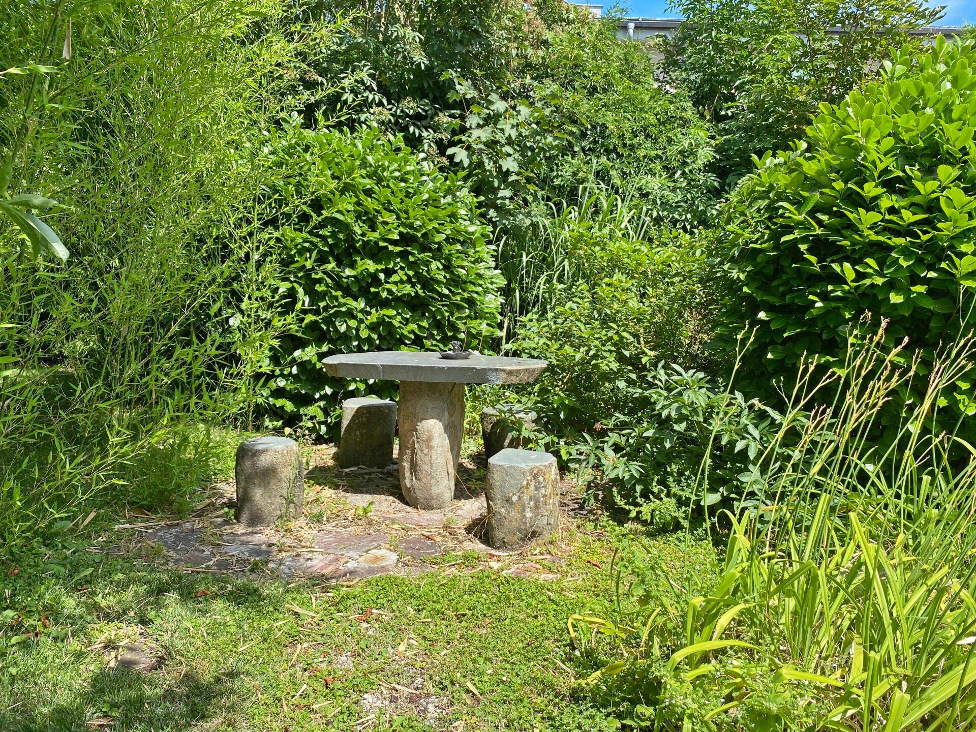 Rustikaler Sitzbereich im Garten mit Steintisch und Stühlen, umgeben von Grün.