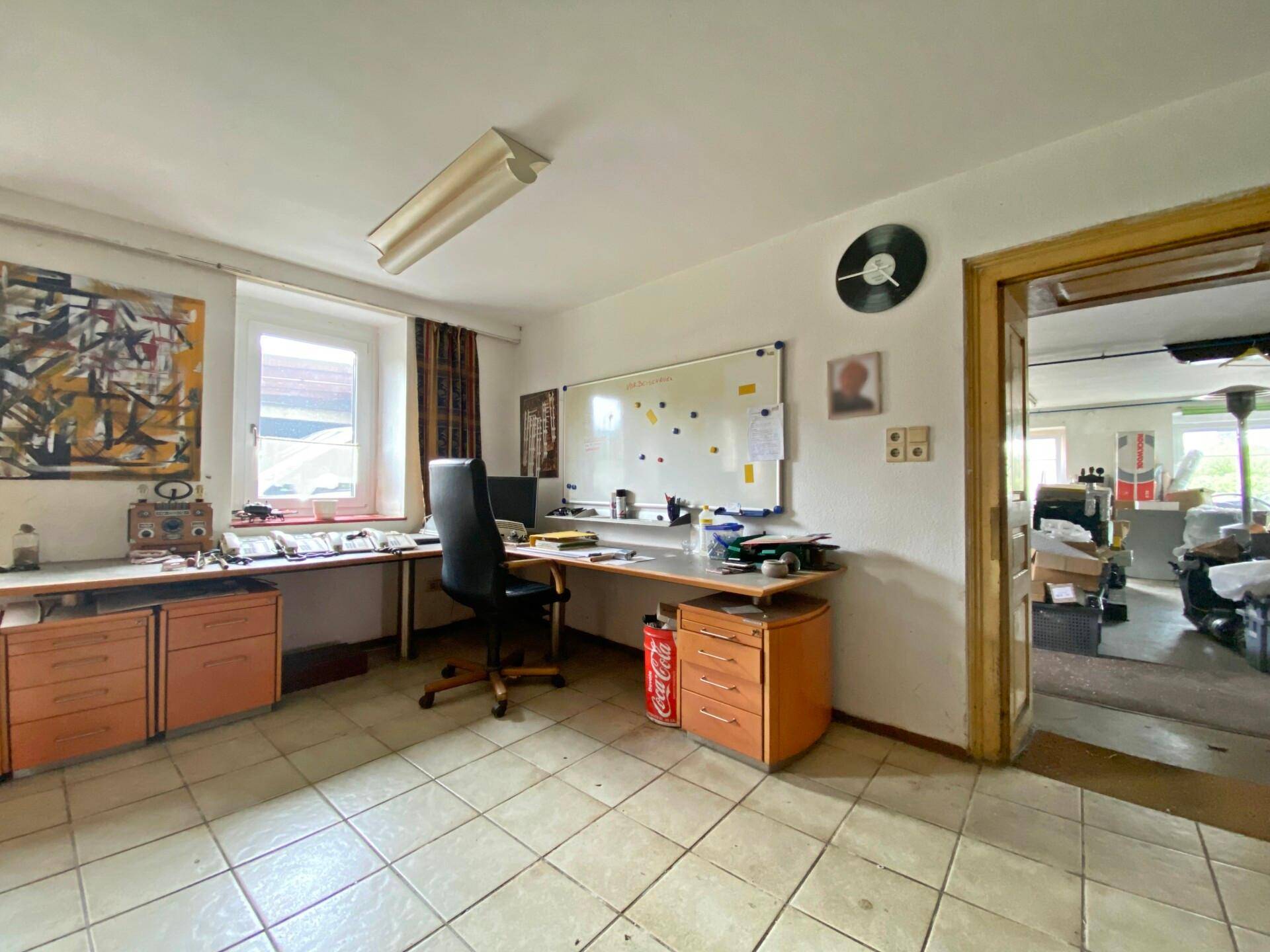 Büro mit Fliesenboden, Schreibtischen und Fenster, angrenzend an einen Lagerraum.