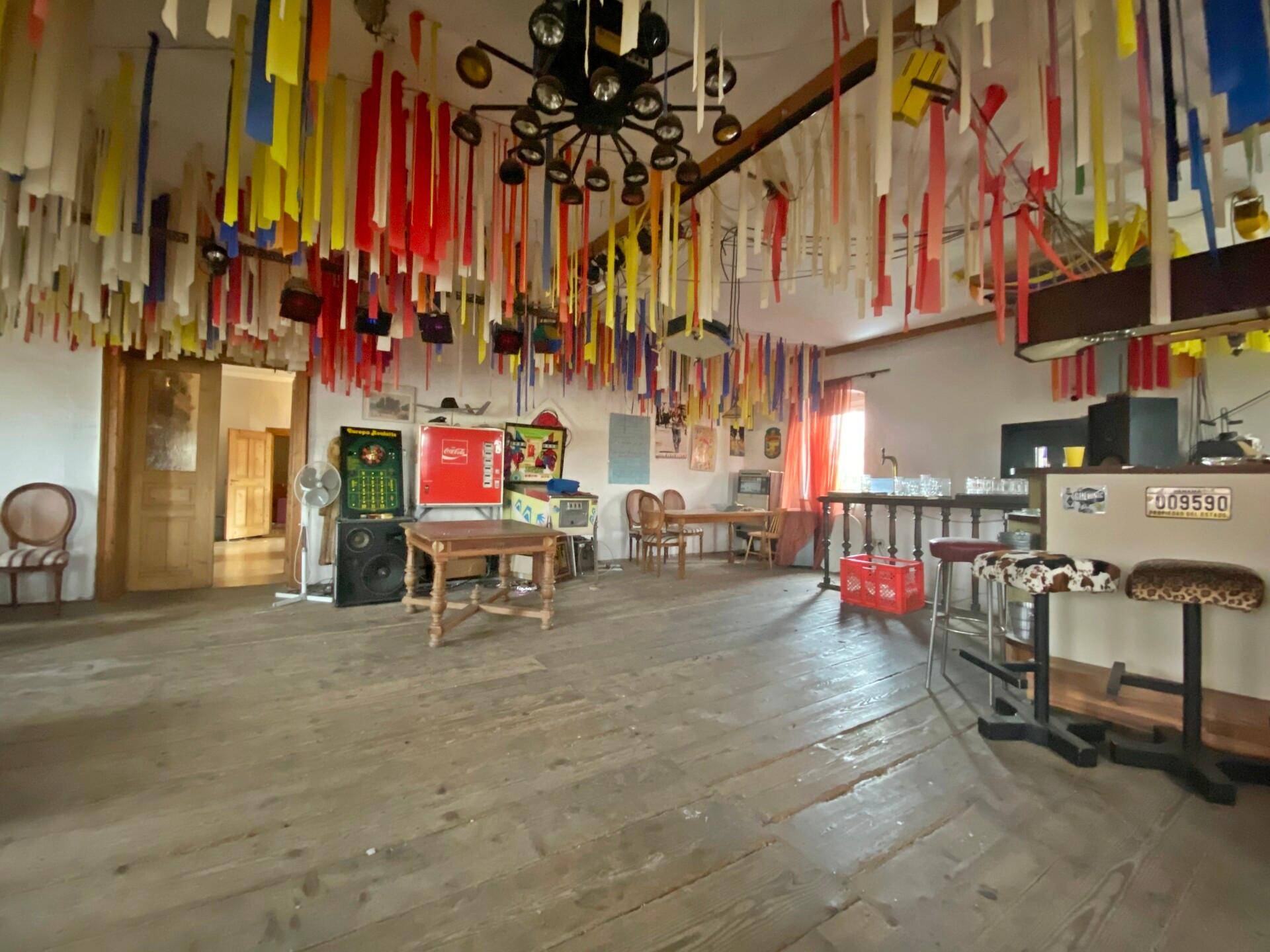 Großer Partyraum mit Holzboden, Bar und bunter Deckenverzierung.