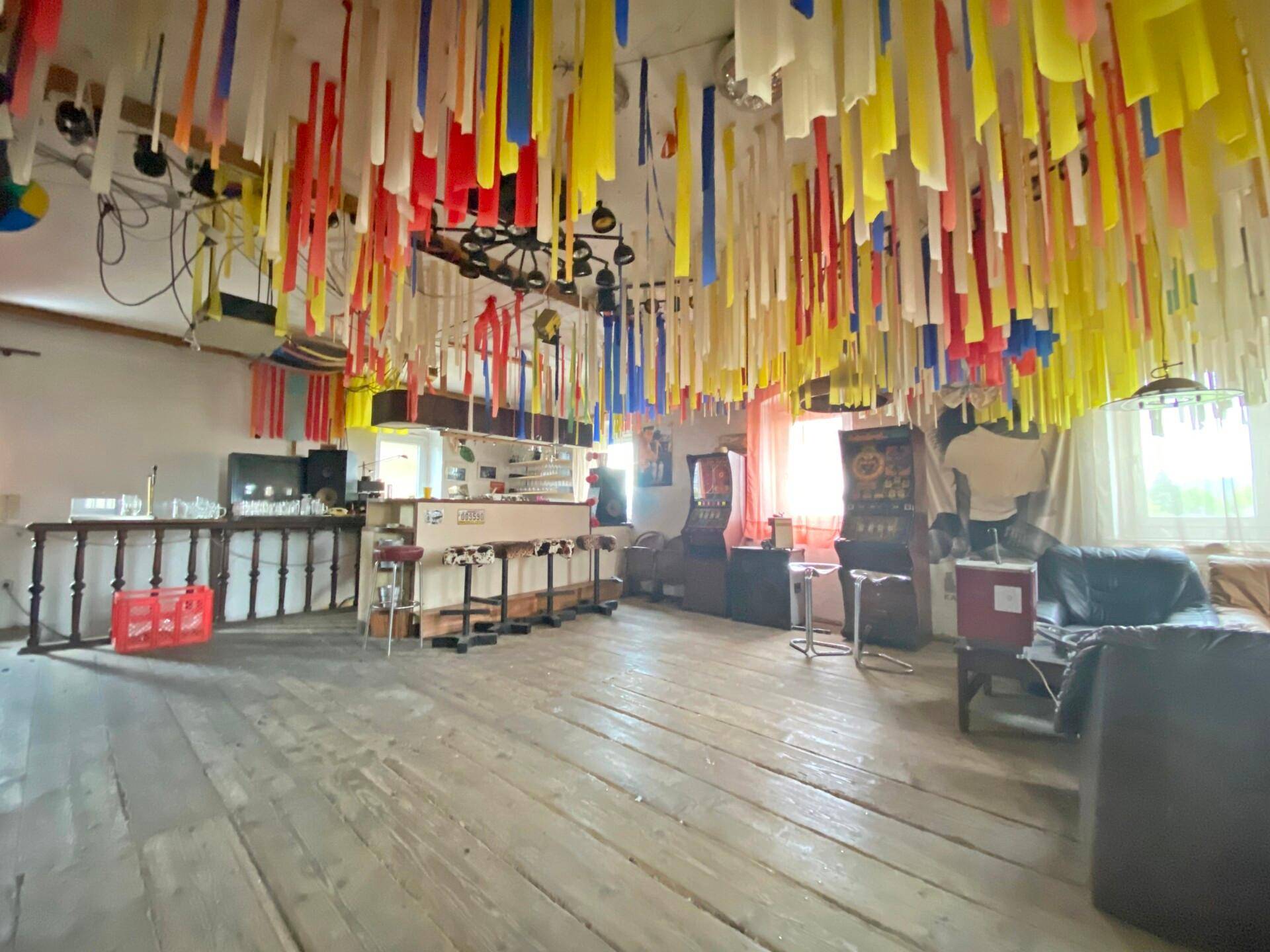 Großer Partyraum mit Holzboden, Bar und bunter Deckenverzierung.