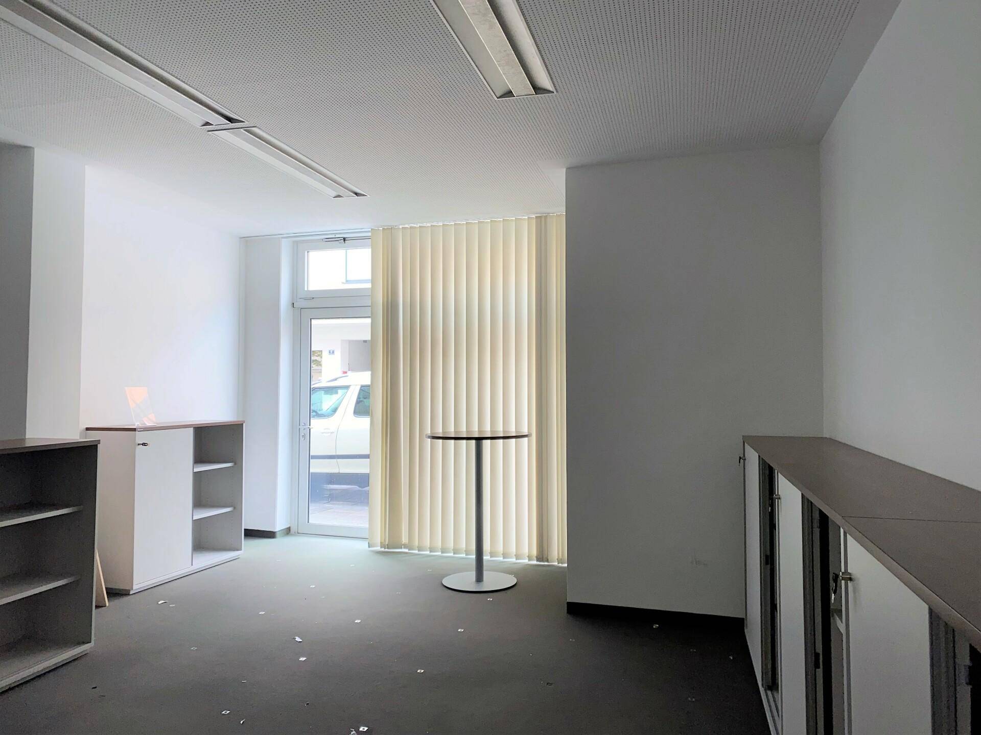 Leerer Büroraum mit dunklem Teppichboden, vertikalen Jalousien an der Tür und einem kleinen Stehtisch.