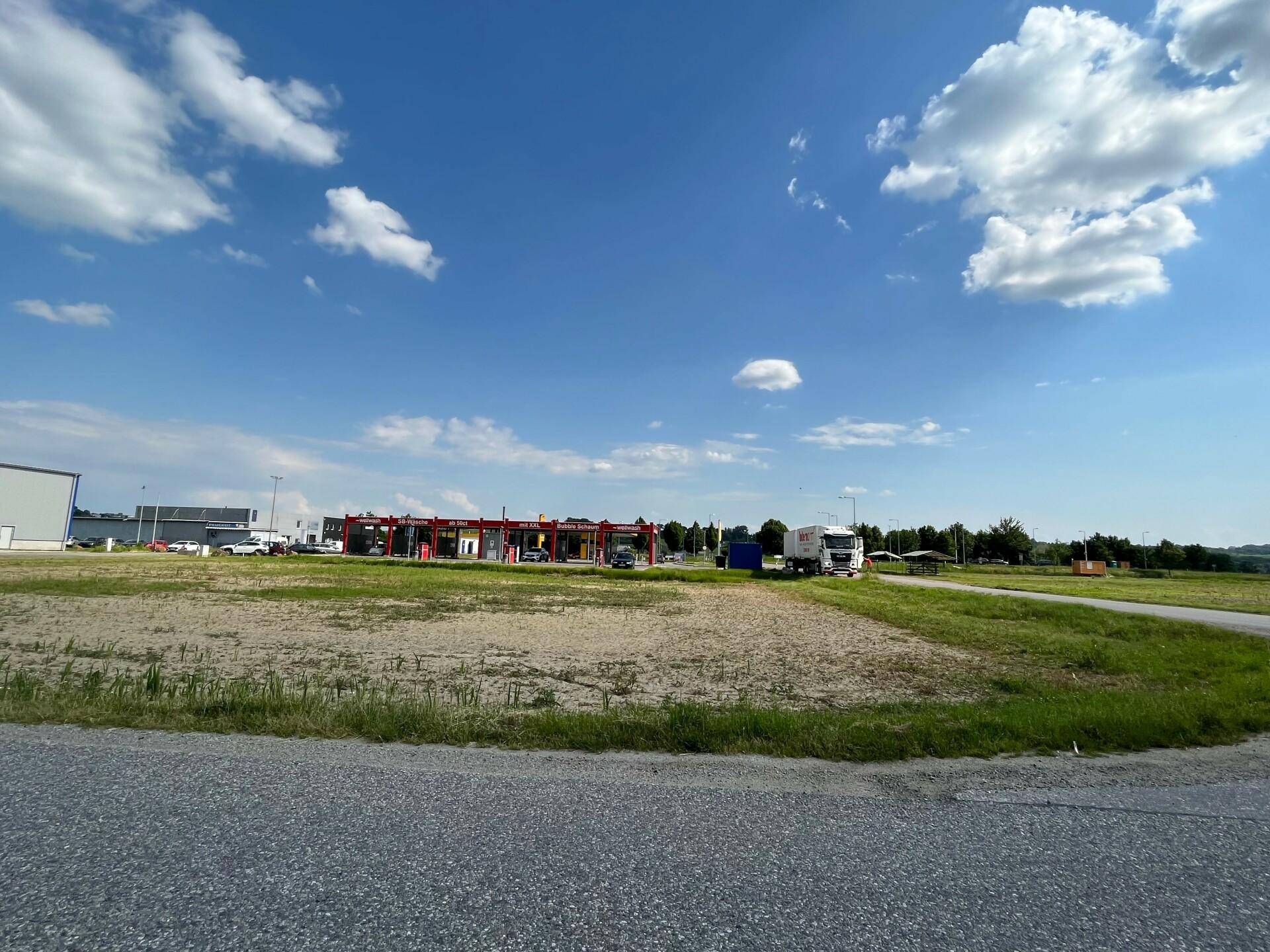 Weitläufiges, unbebautes Grundstück mit Schotter und Gras, angrenzend an eine Straße und Gewerbegebäude.